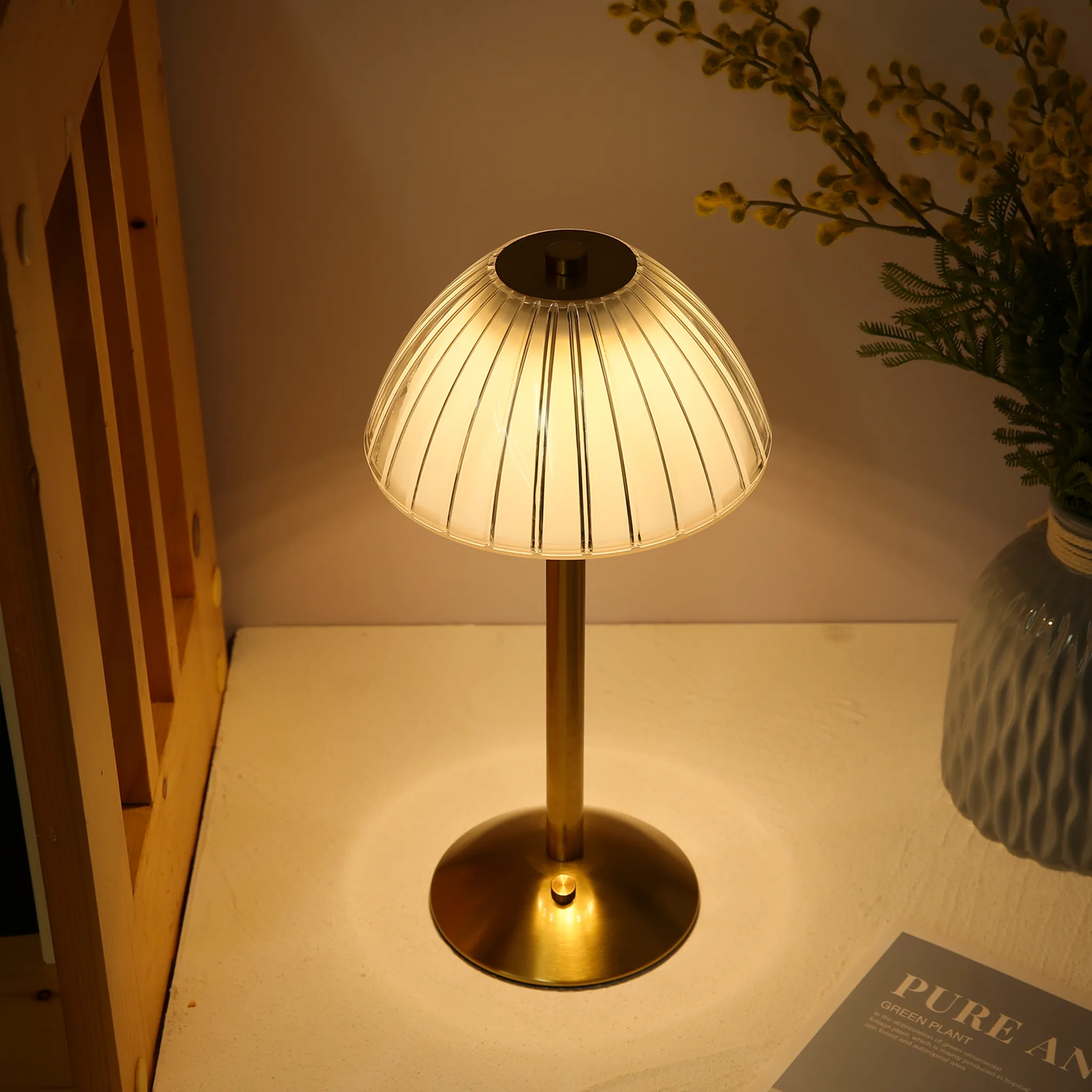 Eine goldene Lampe mit geripptem Glas steht auf einem Tisch neben einem Buch und Pflanzen.
