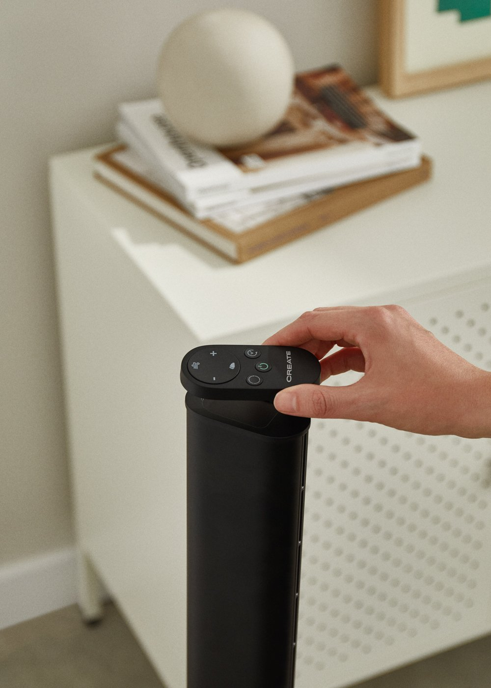 Una mano sostiene un control remoto negro sobre un ventilador negro. Mueble blanco al fondo.