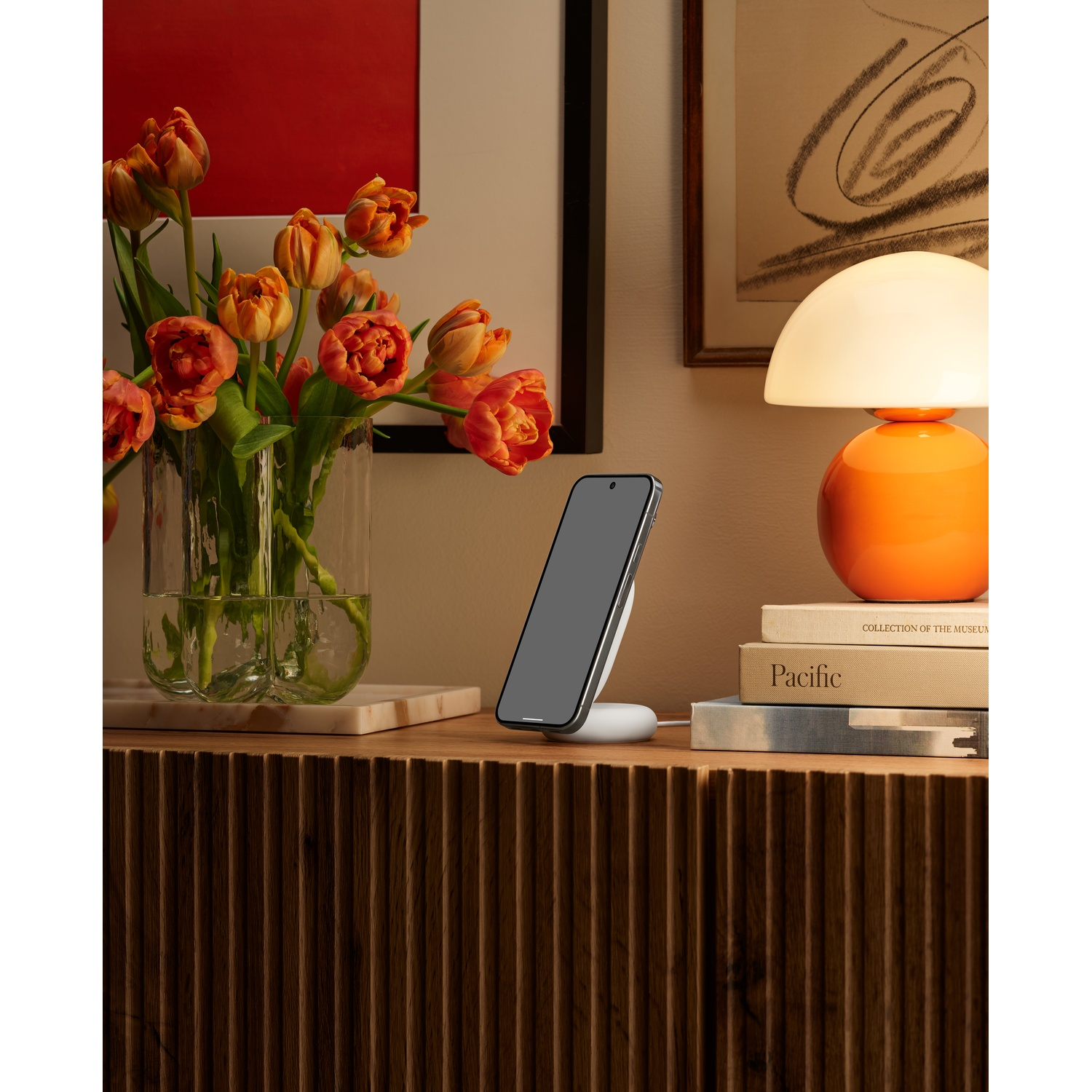 Ein Smartphone lädt auf einem weißen Ständer neben Tulpen und einer orangefarbenen Lampe.