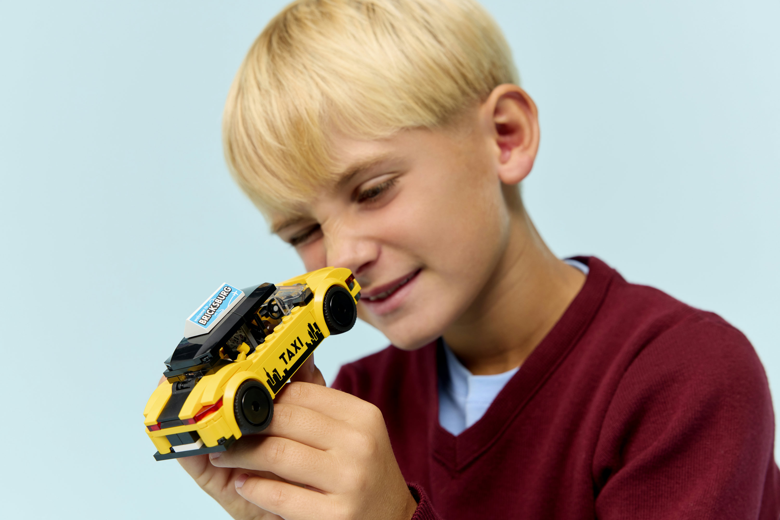 Chłopiec trzyma żółtą taksówkę Lego. Tło jest jasnoniebieskie.