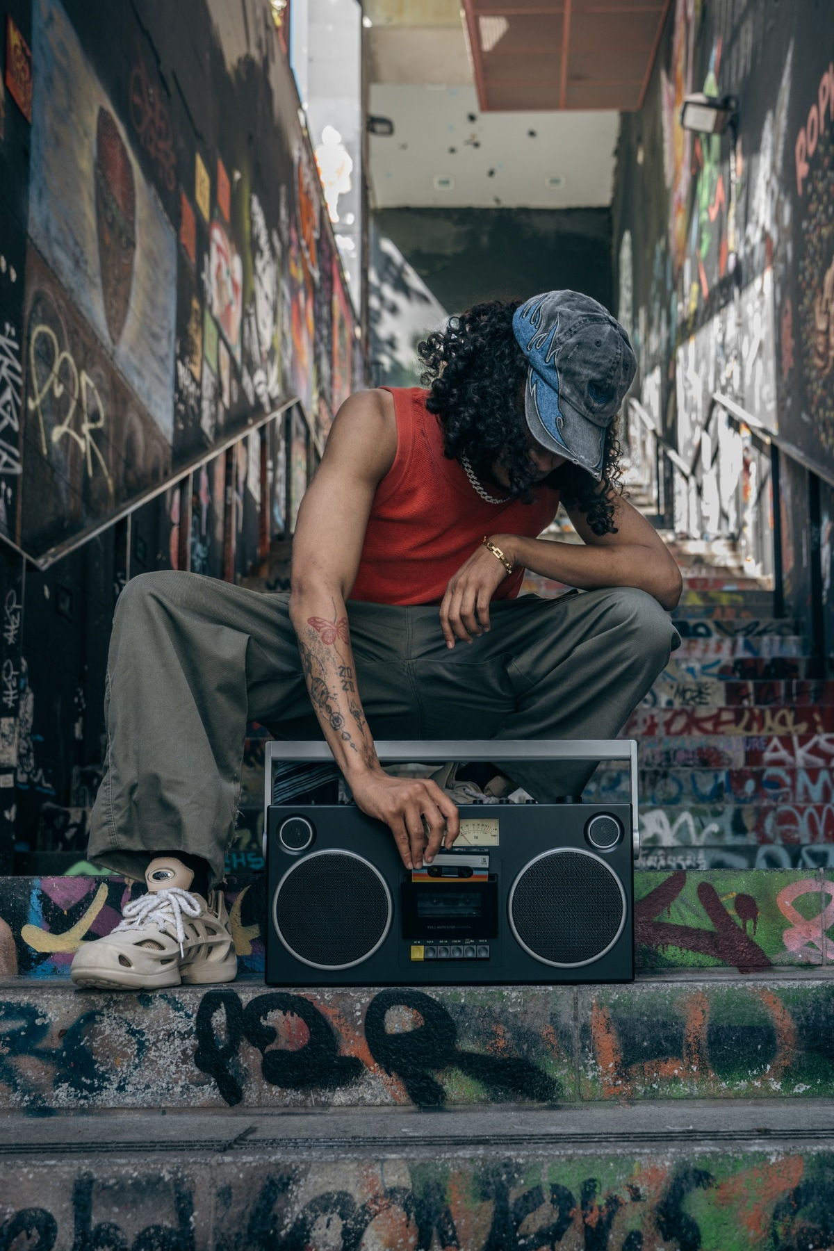Ein Mann mit lockigen Haaren und rotem Tanktop hockt auf einer Treppe, die mit Graffiti bedeckt ist.