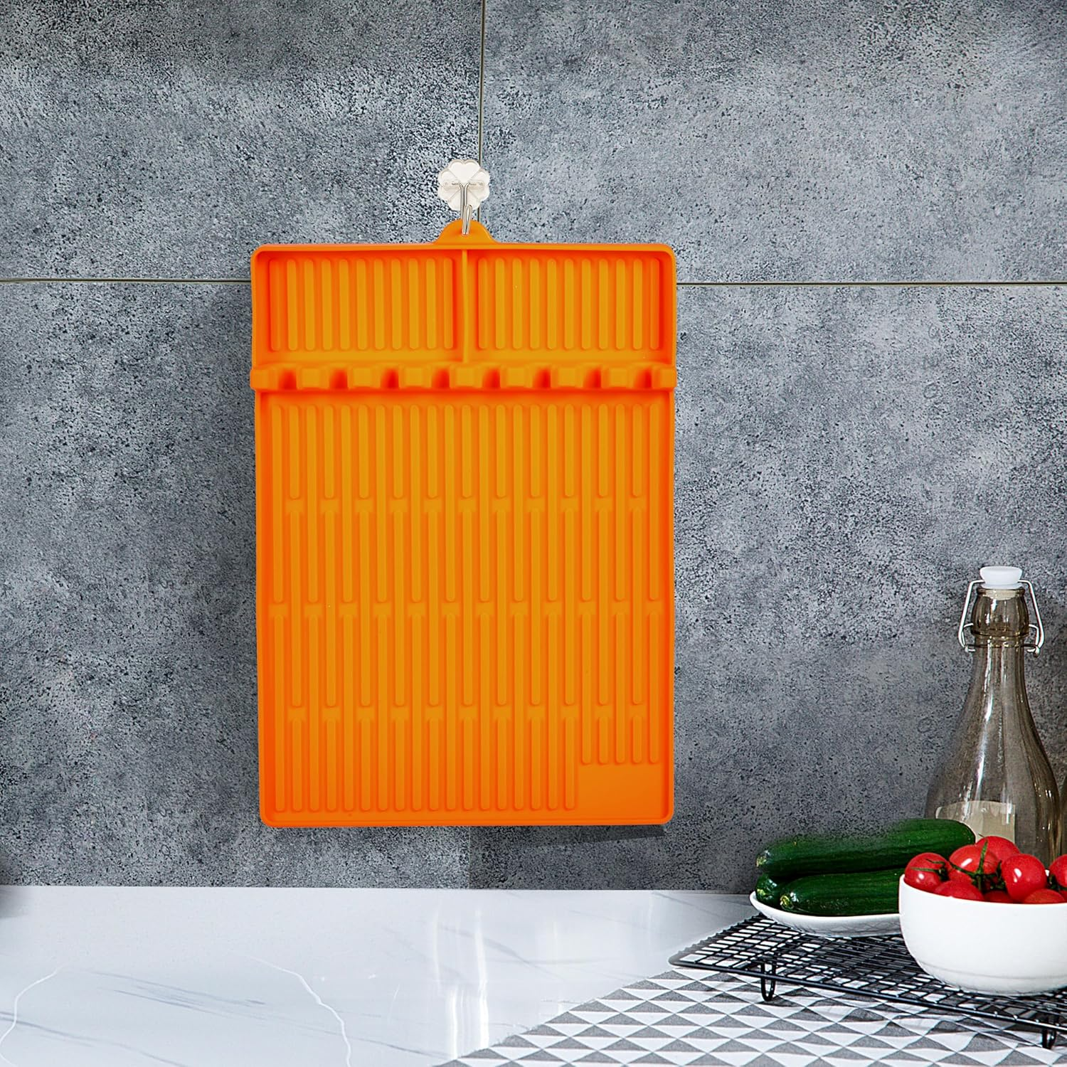 Ein orangefarbenes Abtropfgestell hängt an einer Wand mit grauen Fliesen. daneben sind Gurken und Tomaten.