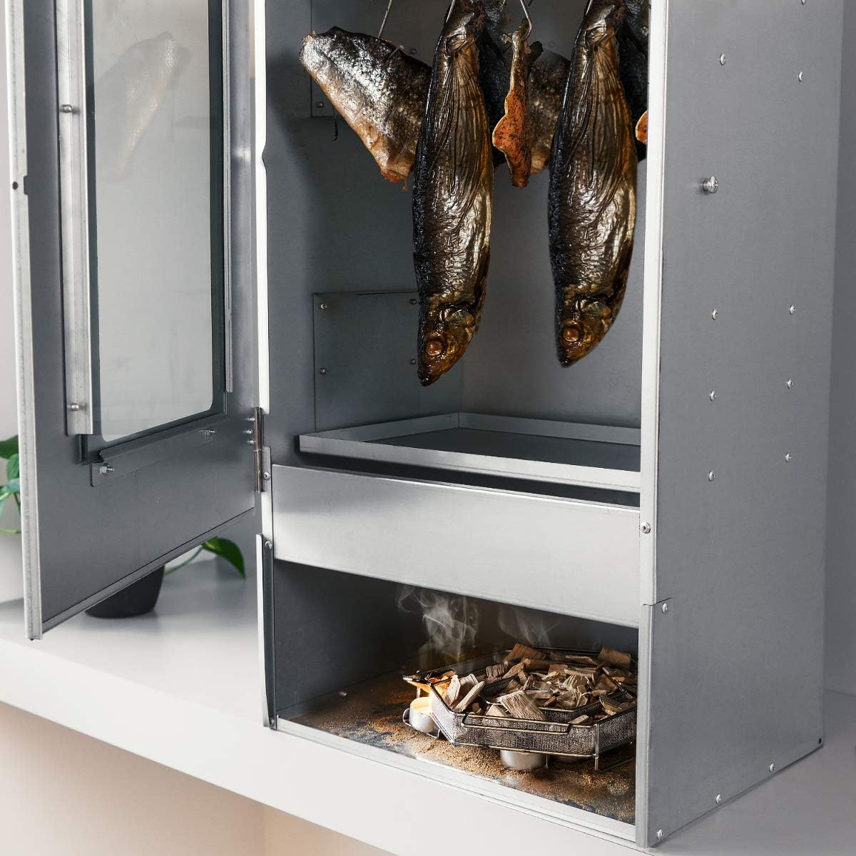 Geräucherte Fische hängen in einem Metallschrank über einer Raucherbox mit Holzspänen.