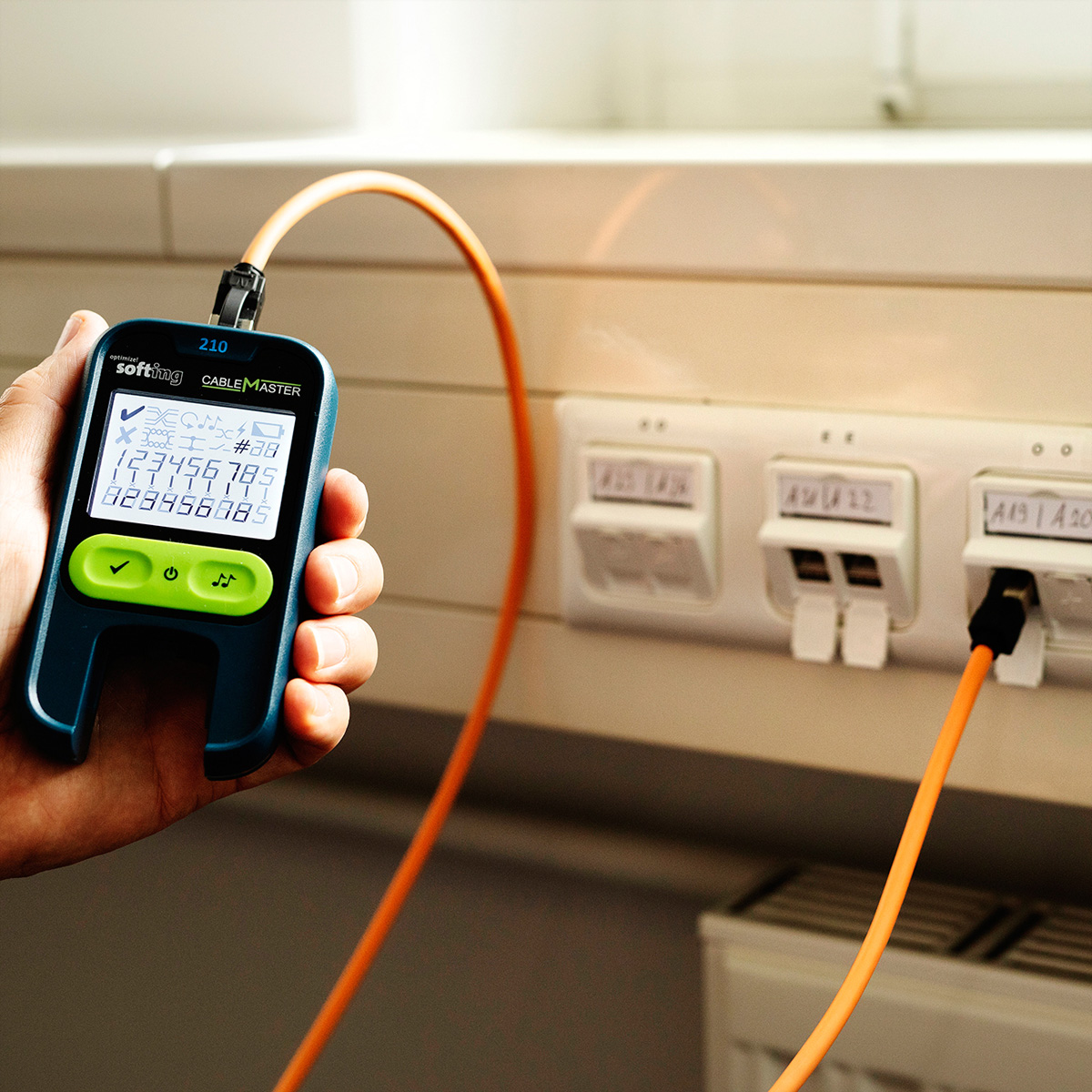 Ein Handheld-Kabeltester mit orangefarbenem Kabel ist mit einer Wandbuchse verbunden.