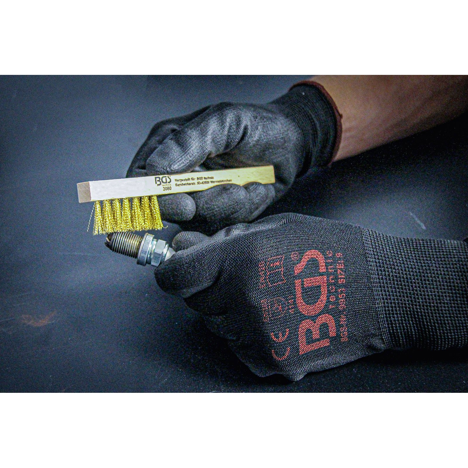 Hände in schwarzen Handschuhen reinigen eine Zündkerze mit einer Messingdrahtbürste.