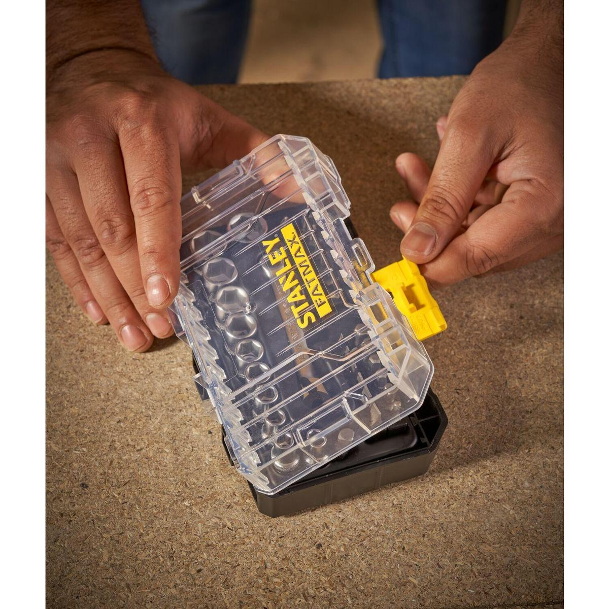 Hände öffnen eine transparente Stanley Fatmax-Box mit Steckschlüsseln auf einer Holzoberfläche.