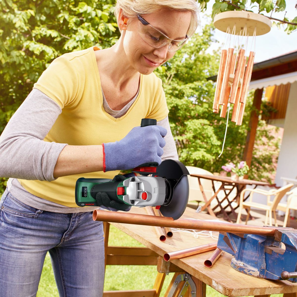 Frau mit Schutzbrille bearbeitet Kupferrohre mit Winkelschleifer im Garten.
