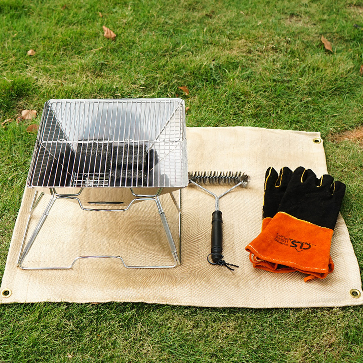 Ein tragbarer Grill, Grillbürste und Handschuhe liegen auf einer Matte im Gras.