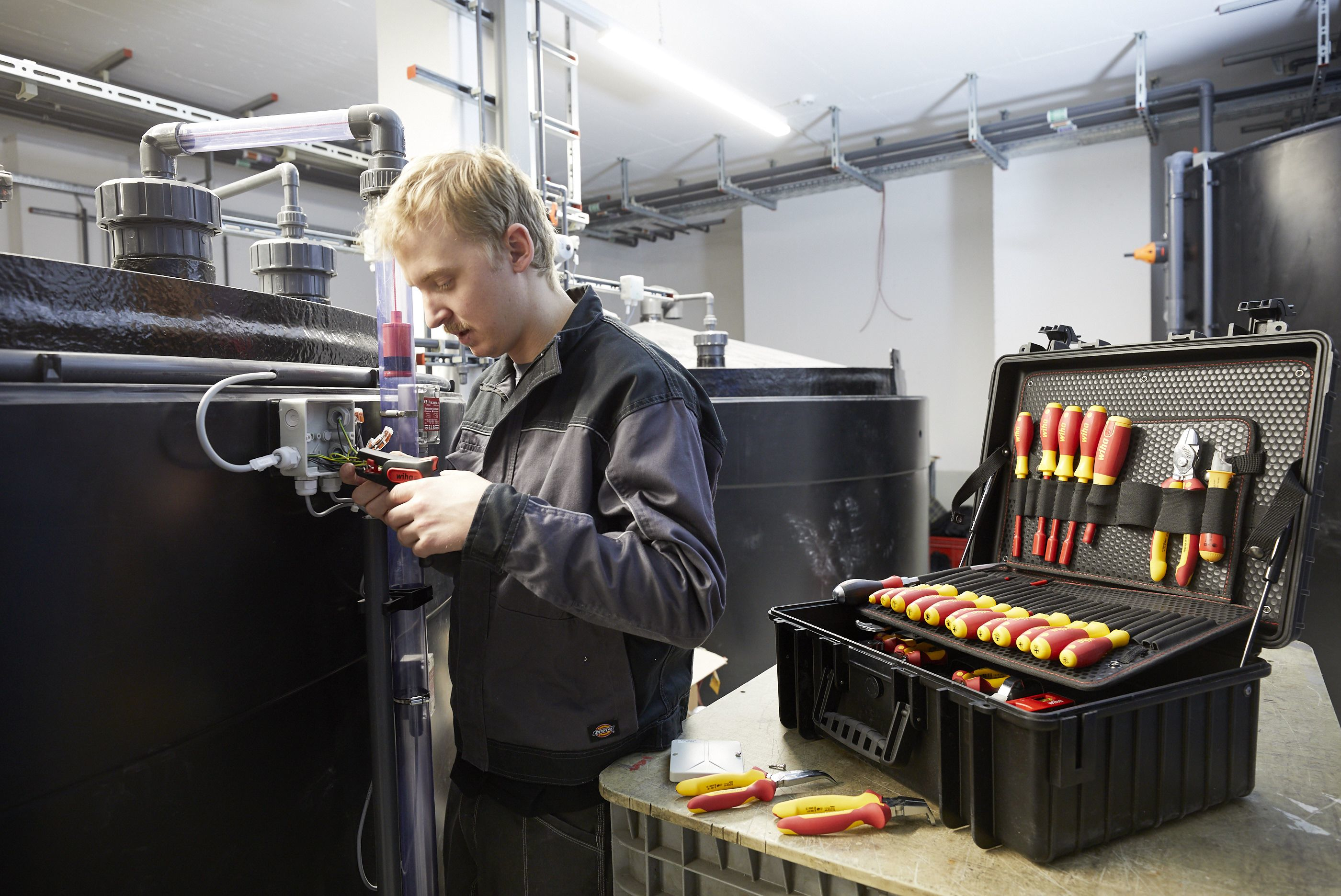 Ein Mann in Arbeitskleidung verdrahtet eine elektrische Anlage neben großen Tanks.