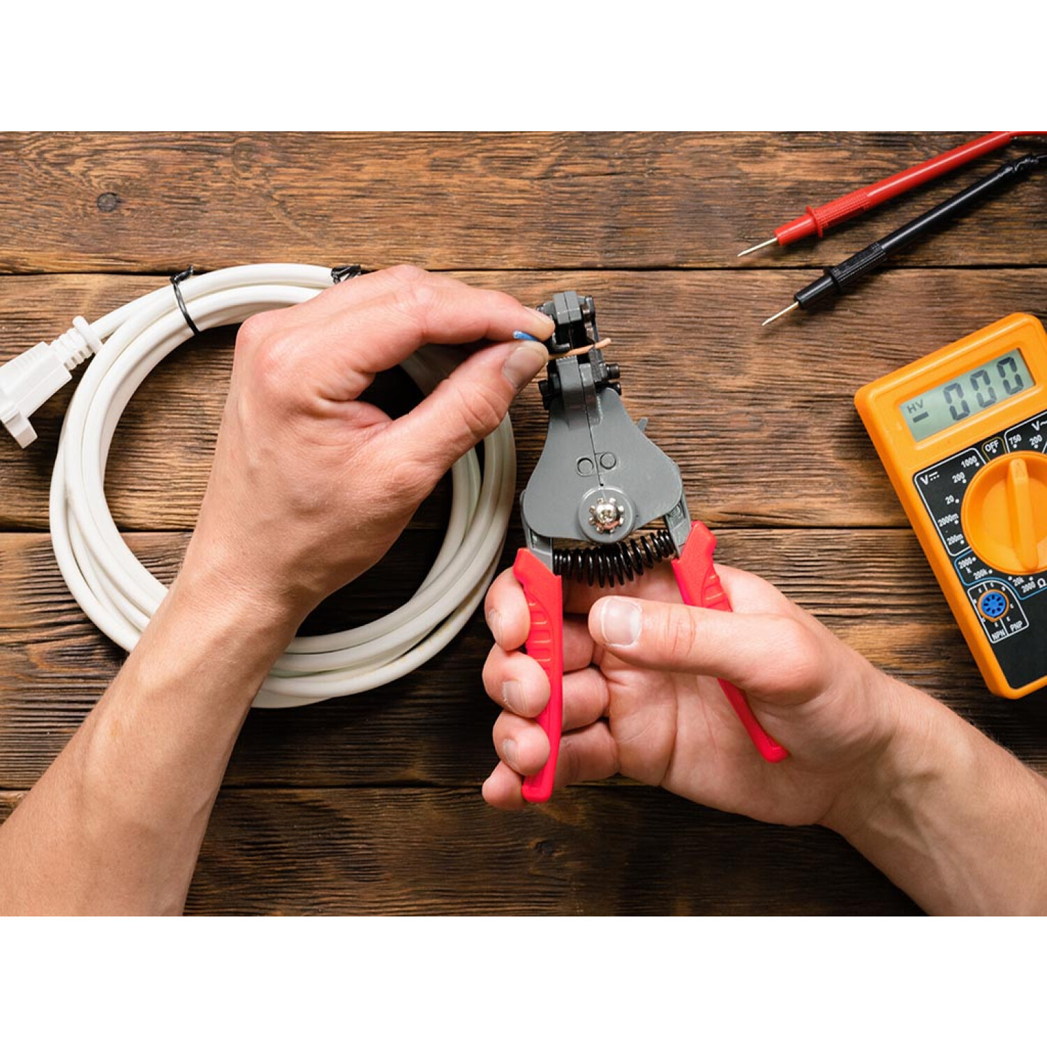 Hände benutzen einen Drahtabisolierer an einem Kabel neben einem Multimeter.