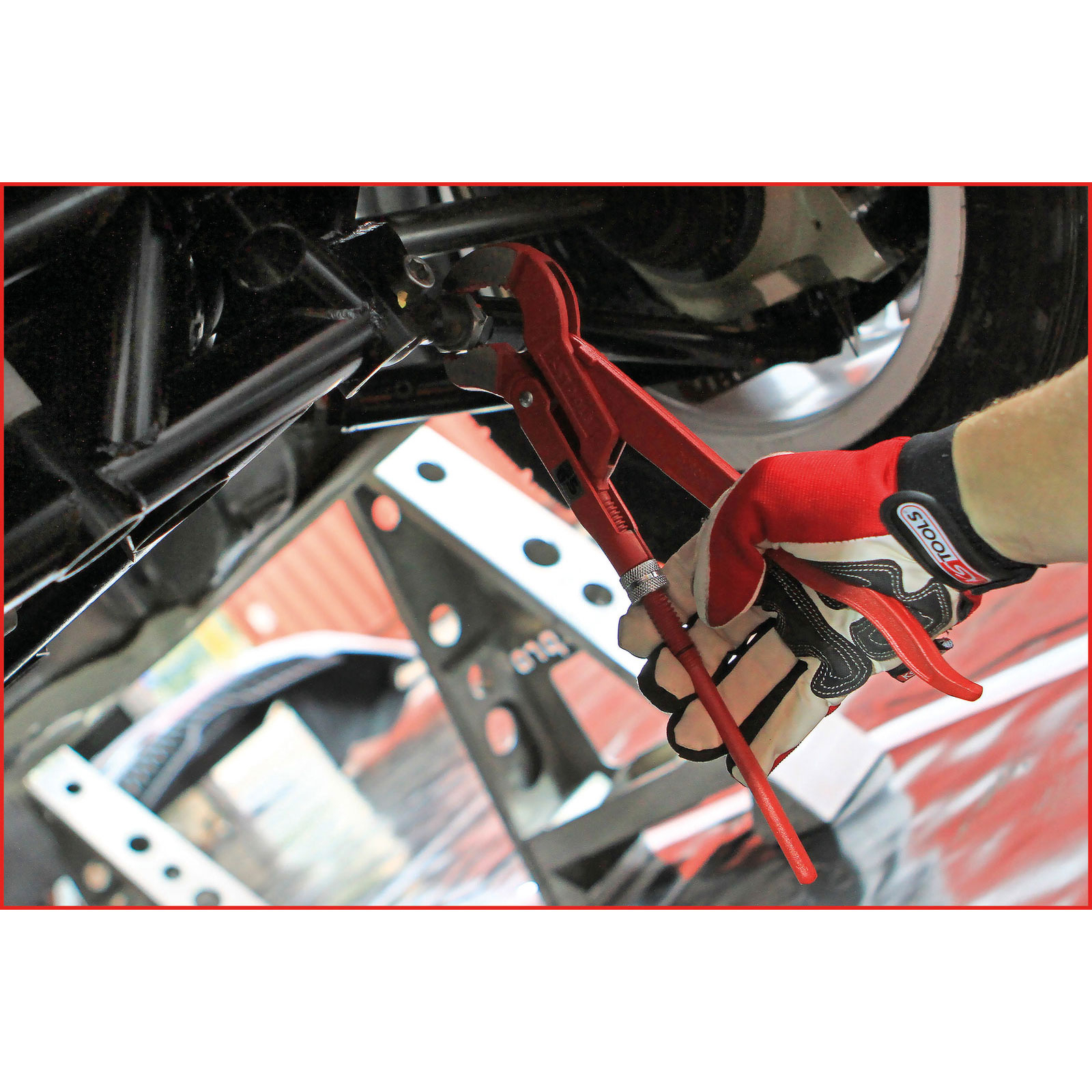 Eine Hand mit rotem Handschuh benutzt eine rote Rohrzange am Fahrwerk eines Autos.