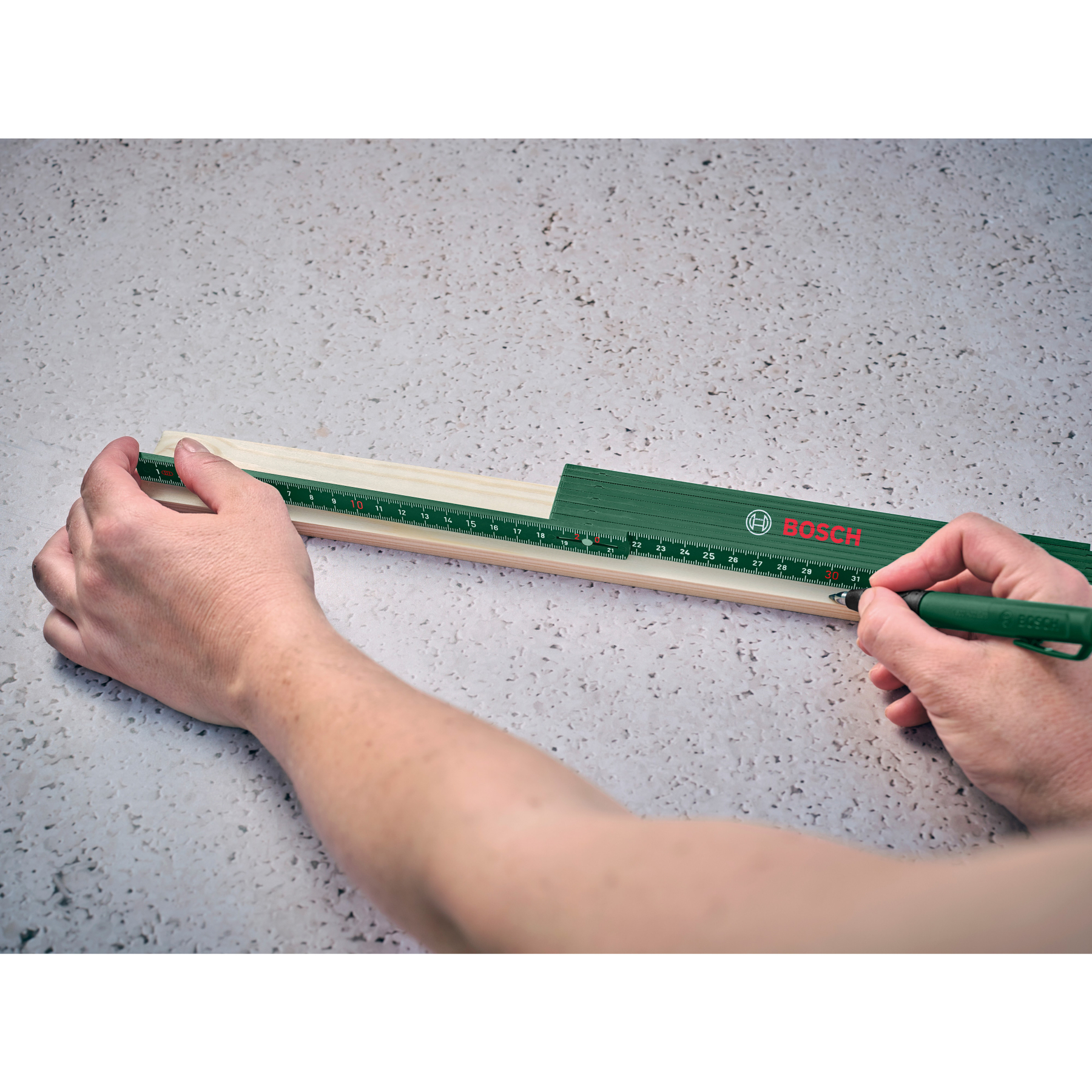 Eine Person misst ein Holzstück mit einem grünen Klapplineal und Bleistift.