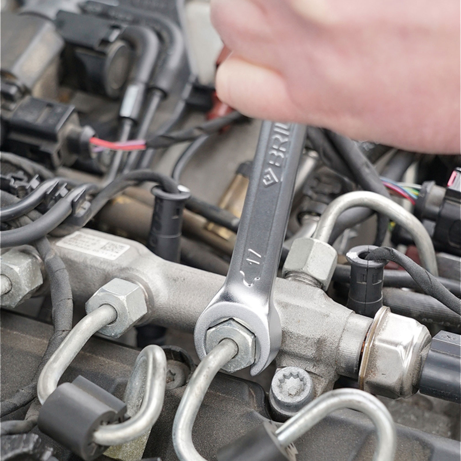 Una mano usa una chiave inglese per stringere un bullone sul rail del carburante di un'auto con fili visibili.