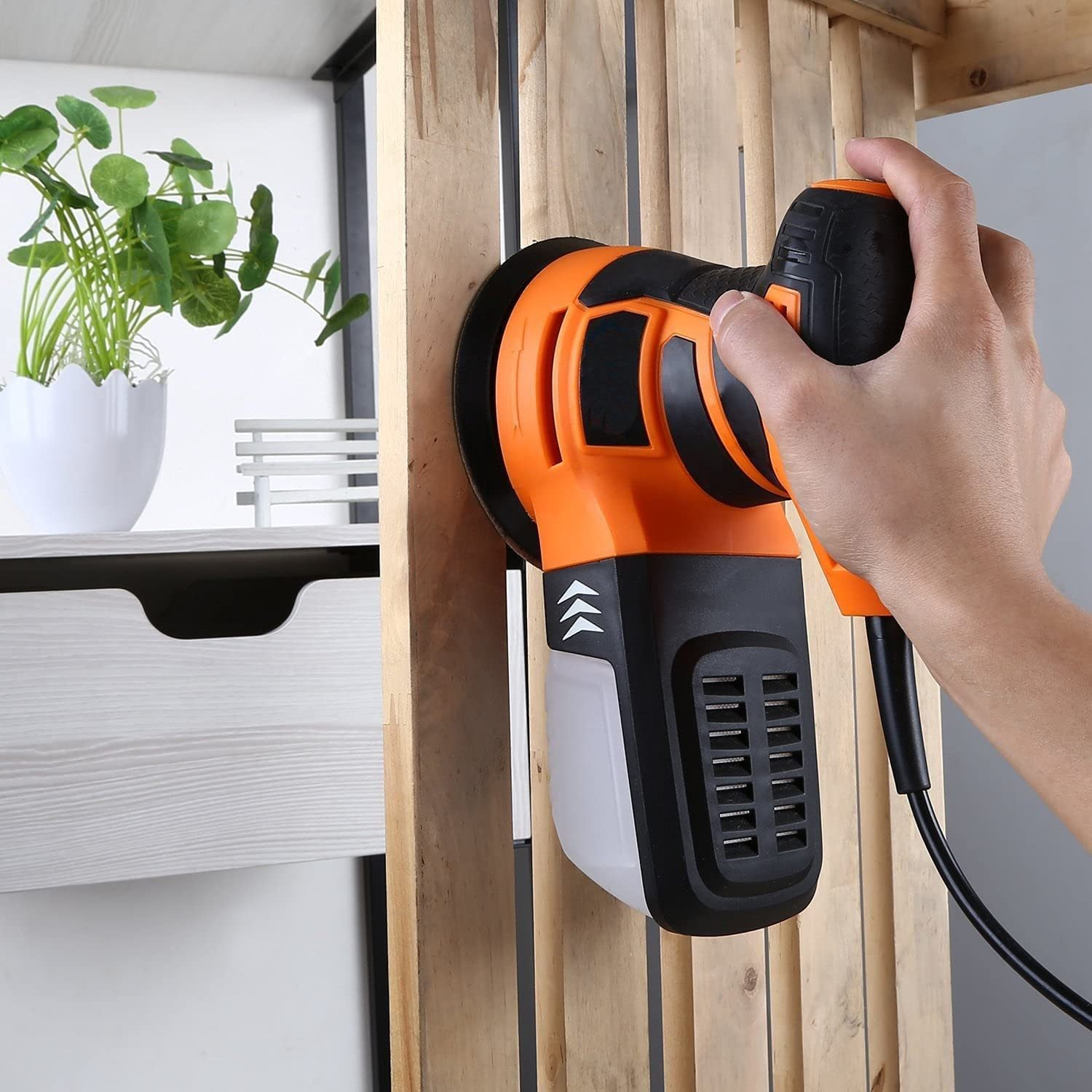 Ein orangefarbener Exzenterschleifer wird von einer Hand gehalten, die ihn auf Holz richtet.