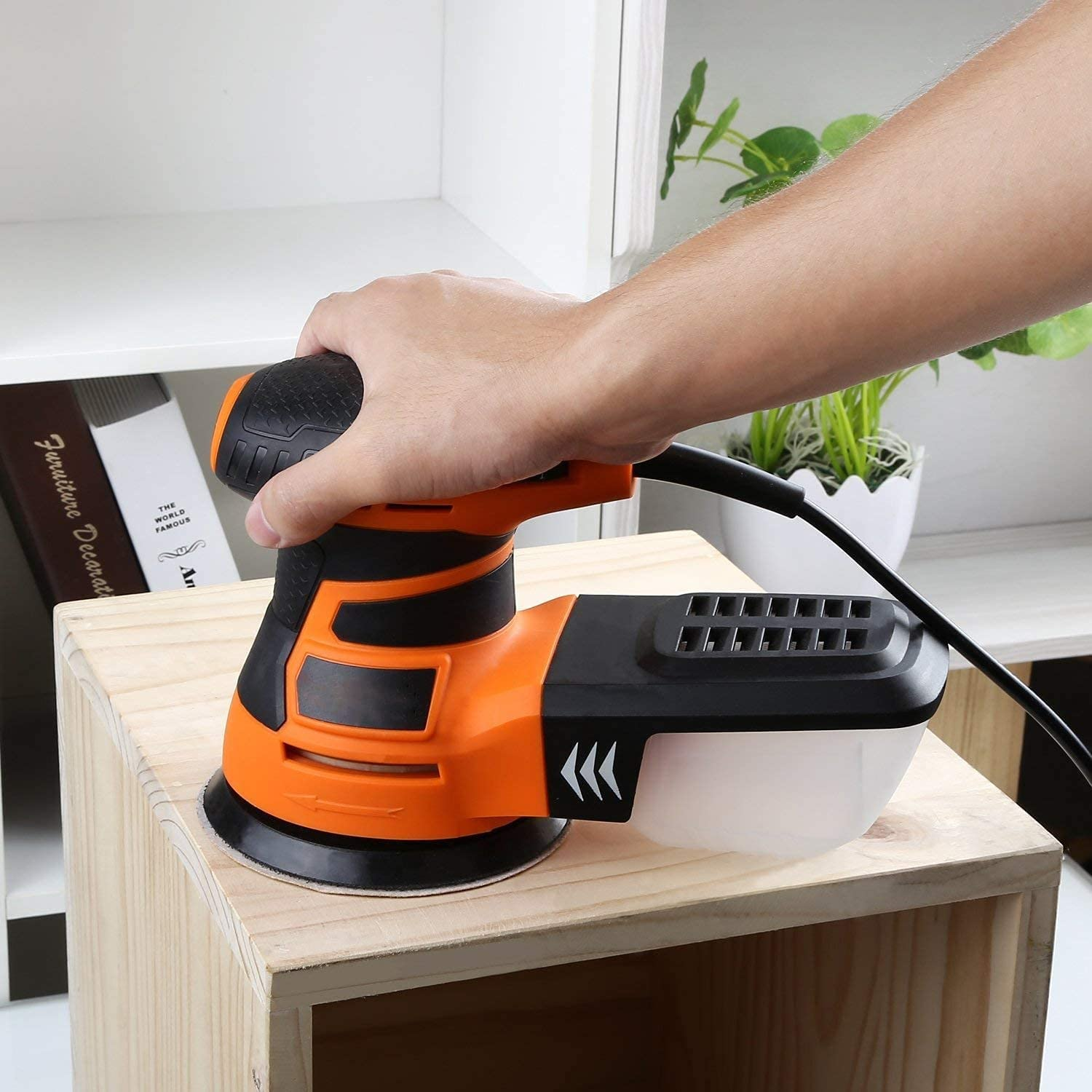 Eine Hand hält einen orangefarbenen Exzenterschleifer auf einem Holzkasten.