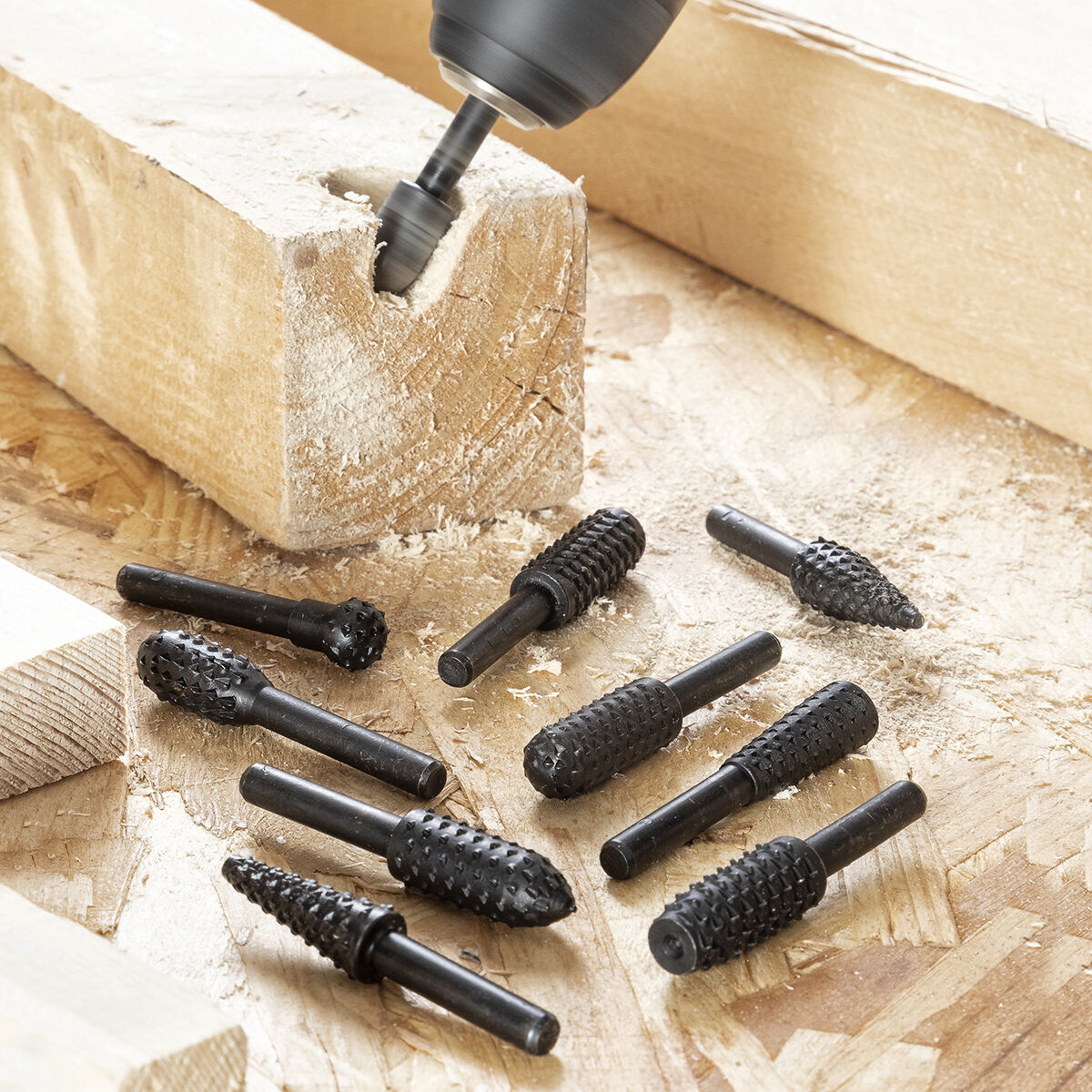 Un trapano lavora un blocco di legno; accanto giacciono diverse frese.