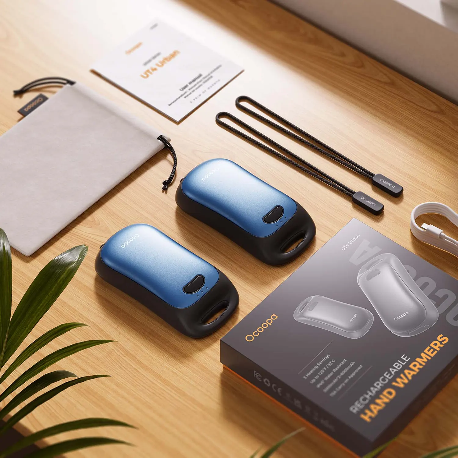 Dwa niebieskie podgrzewacze do rąk Ocoopa na drewnianym biurku z instrukcją, etui i kablem.