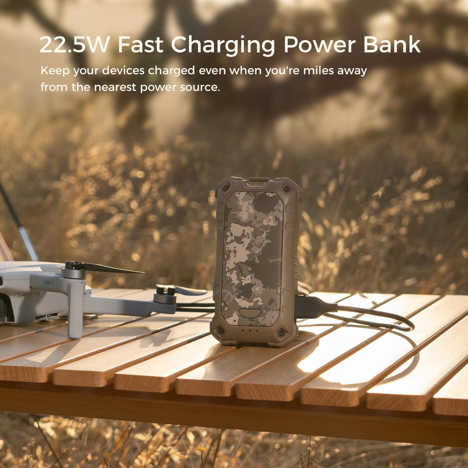 Power bank w kamuflażu ładuje się obok drona DJI Mini 2 na drewnianym stole.