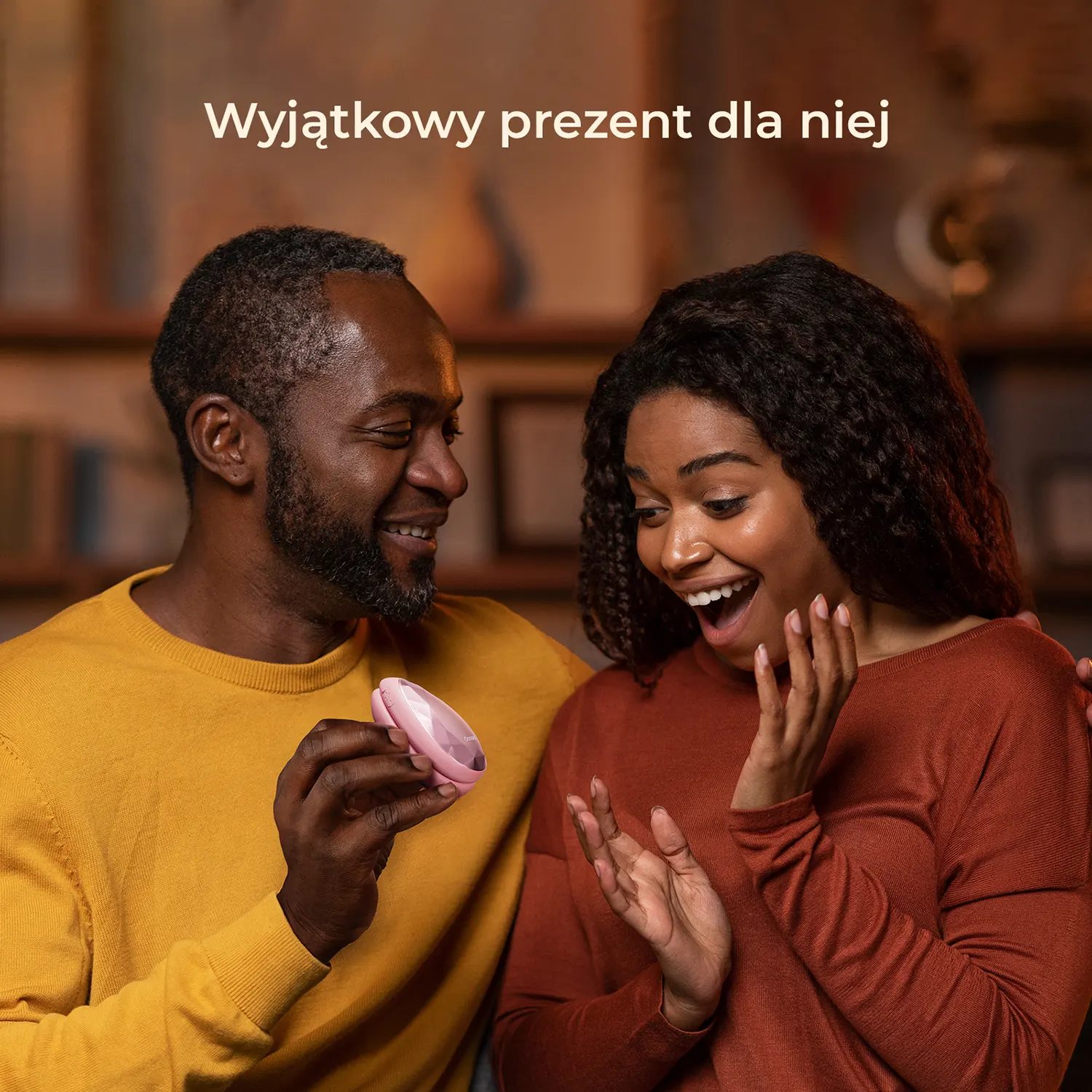 Mężczyzna w żółtym swetrze trzyma różowy prezent dla zaskoczonej kobiety.