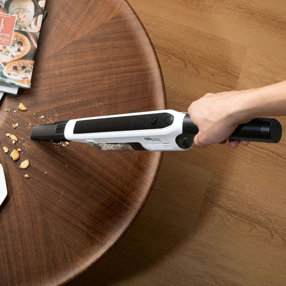 Una mano sostiene una aspiradora blanca recogiendo migas de una mesa de madera.