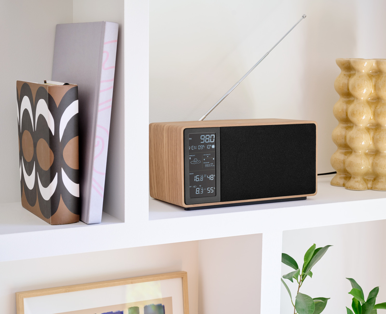 Drewniane radio z czarnym głośnikiem i wyświetlaczem na półce z książkami i wazonem.