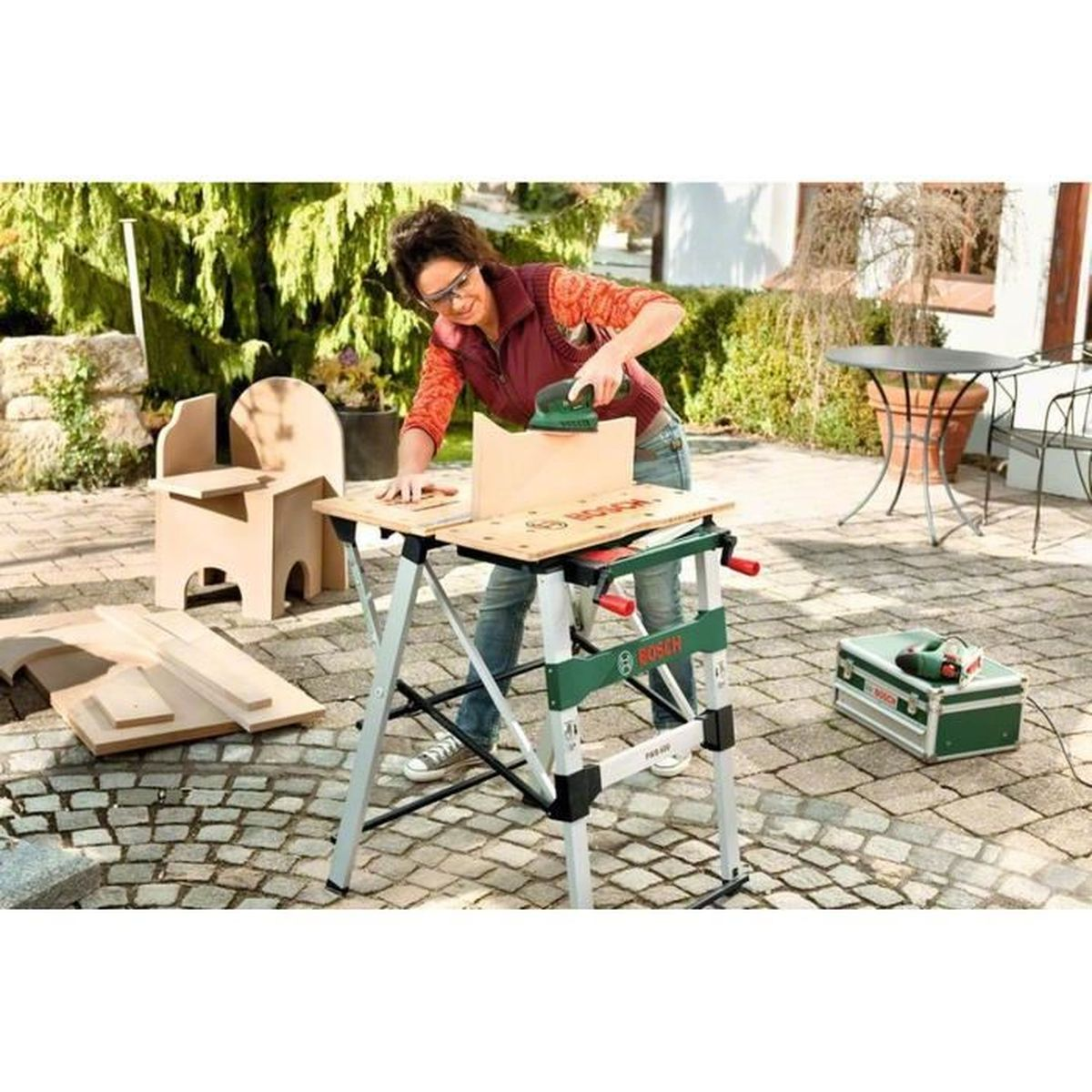Frau bearbeitet Holz auf Werkbank im Garten. Kiste daneben.