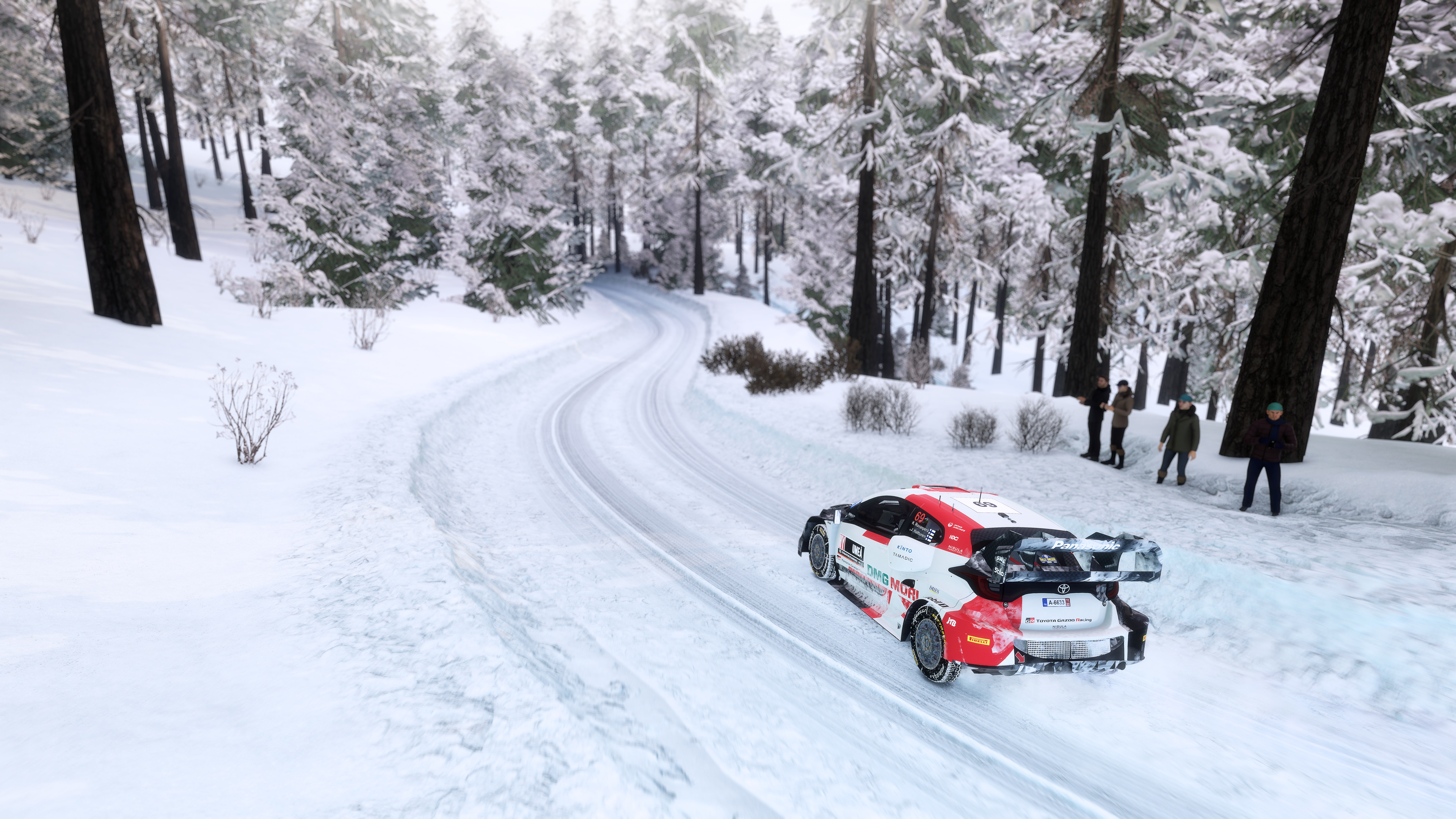 Ein Rallyeauto fährt auf einer verschneiten Straße durch einen Wald. Personen stehen am Wegesrand.