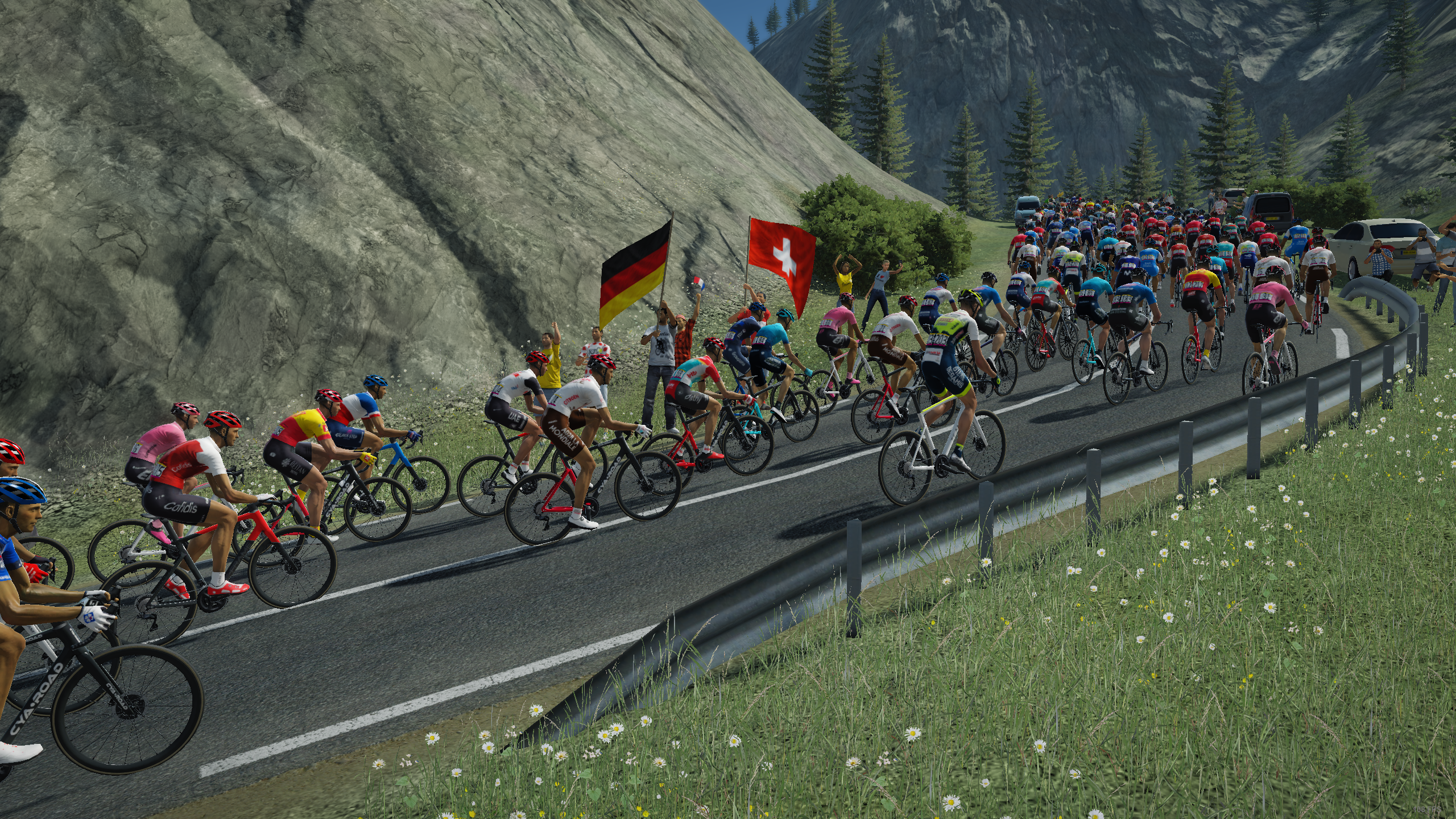 Radsportler fahren bergauf, Zuschauer winken mit Fahnen. Berglandschaft im Hintergrund.