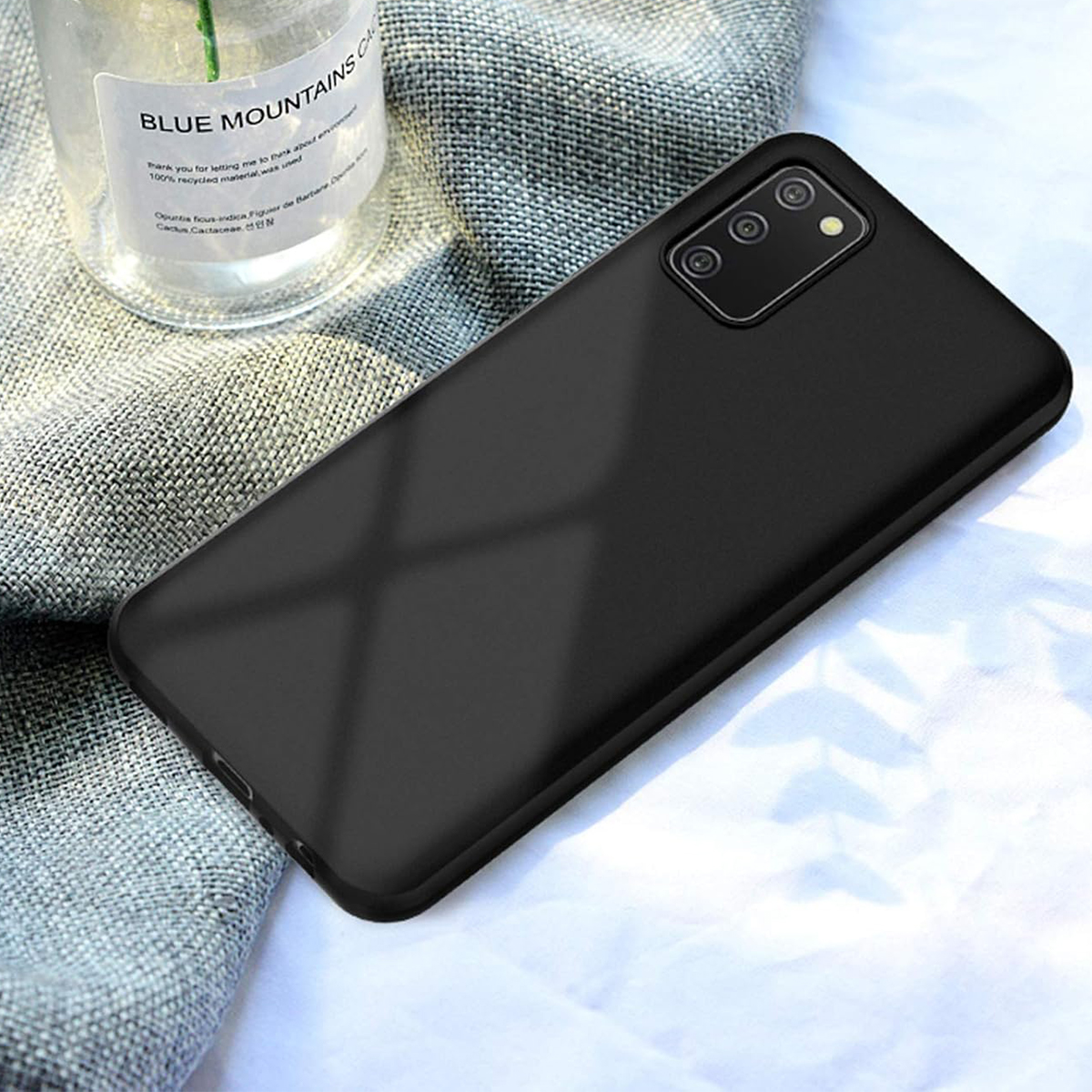 Smartphone nero con tripla fotocamera su tessuto grigio accanto a una bottiglia di vetro.