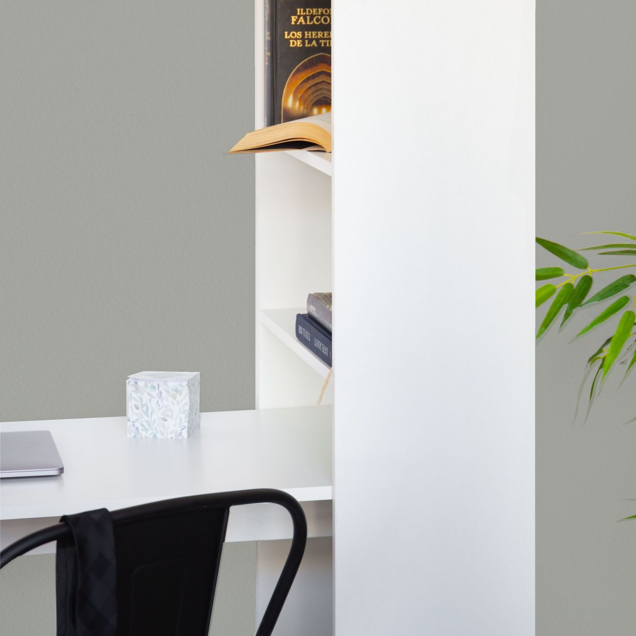 Un escritorio con portátil, libro y rincón de estantería con plantas verdes.