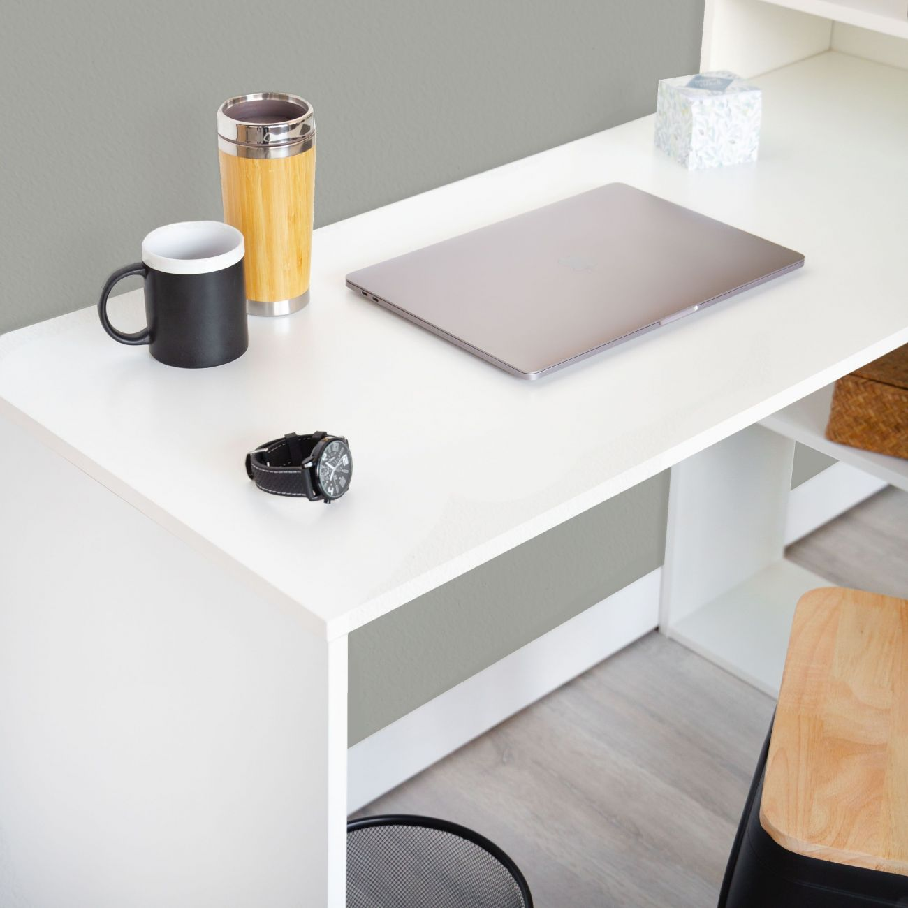 Un escritorio blanco con laptop, taza, termo y reloj. Una silla está cerca.