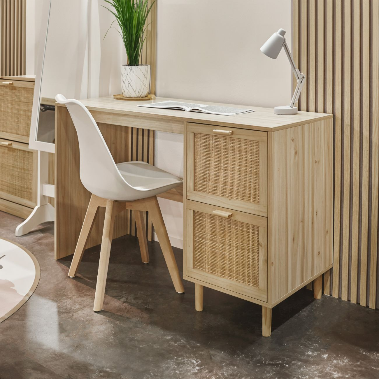 Un escritorio de madera clara con detalles de ratán, silla blanca y lámpara.