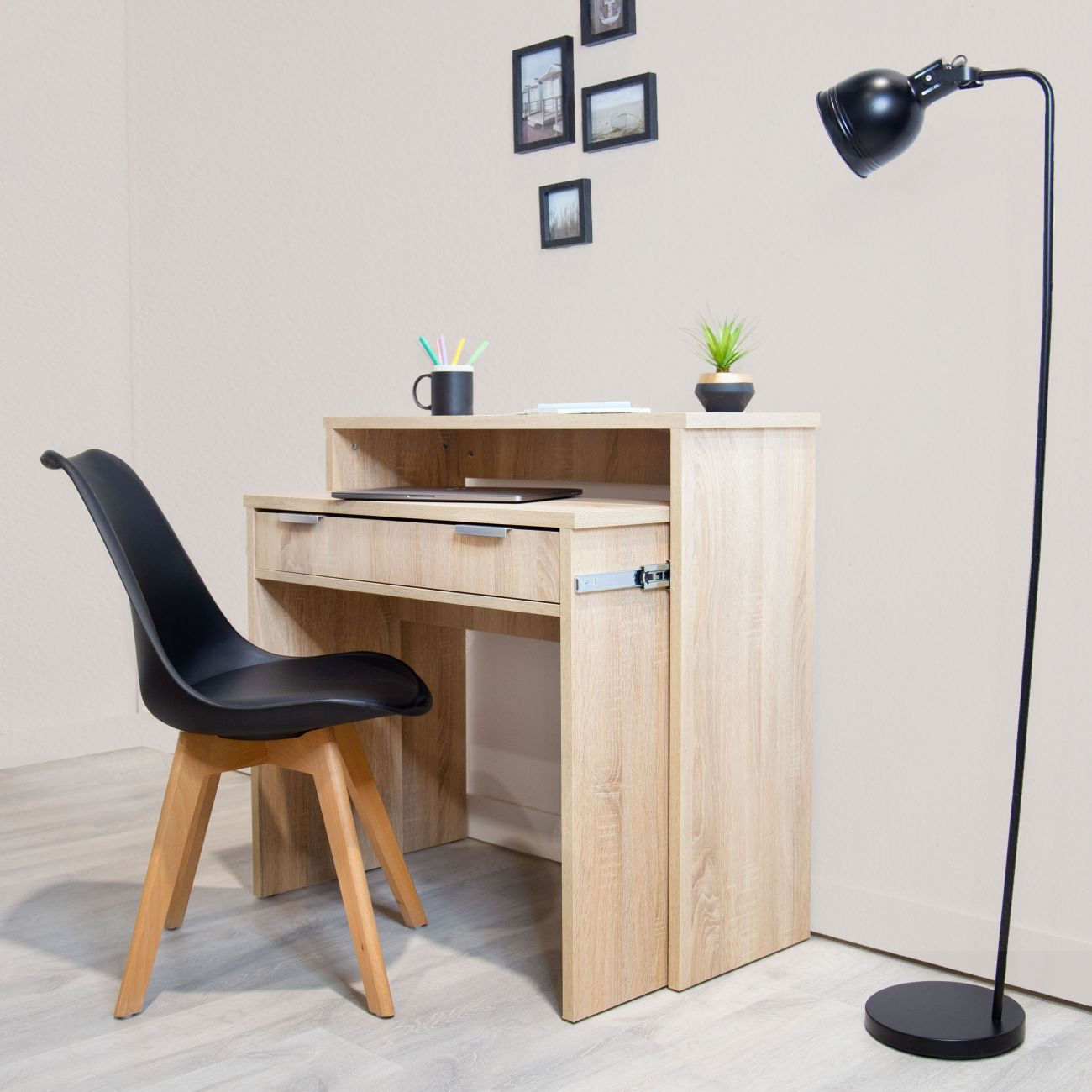 Un escritorio de madera clara con cajones, silla negra y lámpara de pie.