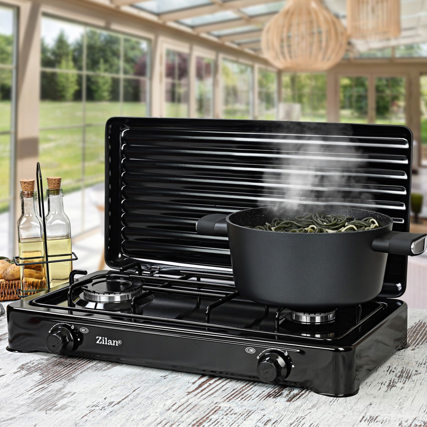 Czarna kuchenka gazowa z garnkiem gotującym parujący makaron, oranżeria w tle.