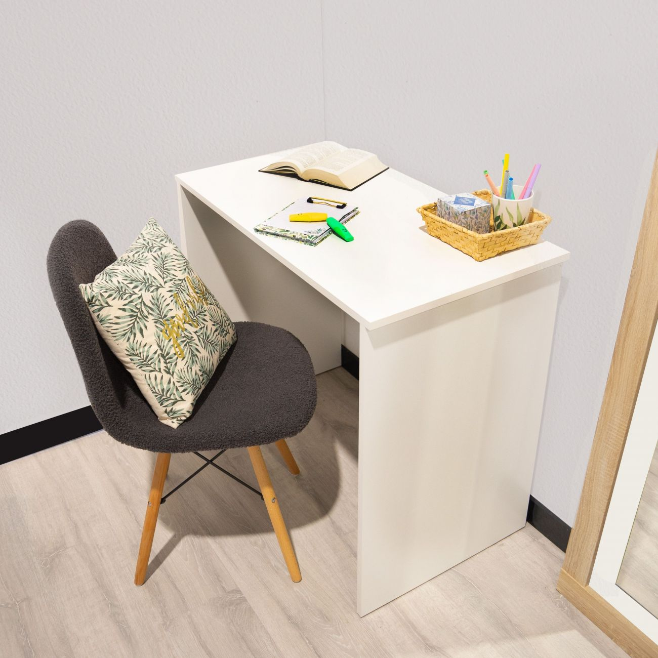 Un escritorio blanco con libro abierto, bolígrafos y cesta. Una silla con cojín está cerca.