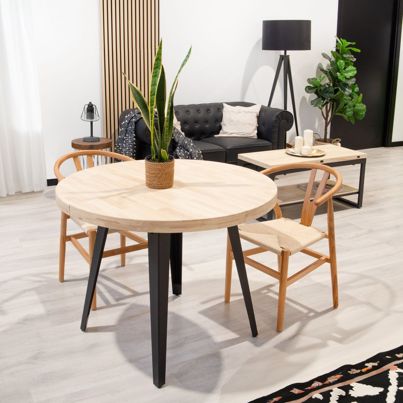 Mesa redonda de madera con patas negras, dos sillas de madera, sofá negro, lámpara.