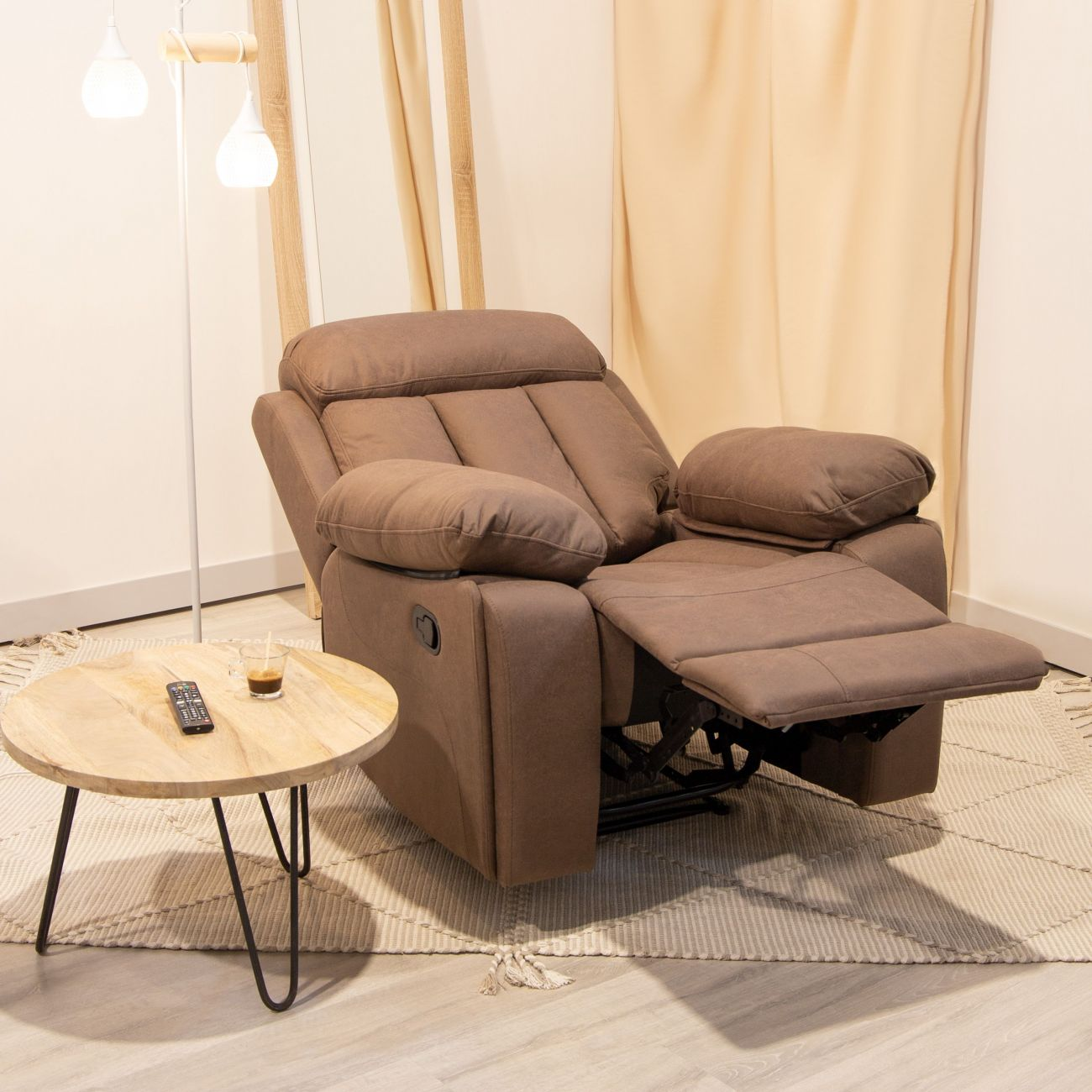 Un sillón reclinable marrón con reposapiés, mesa redonda de madera con mando y lámpara de pie.