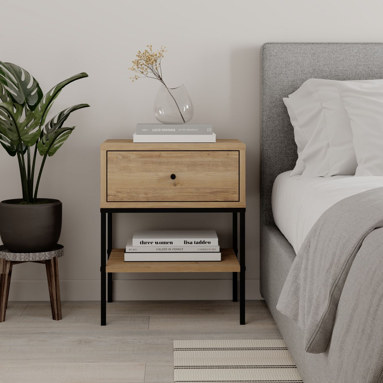 Mesita de noche de madera con cajón, libros, jarrón y planta junto a una cama.