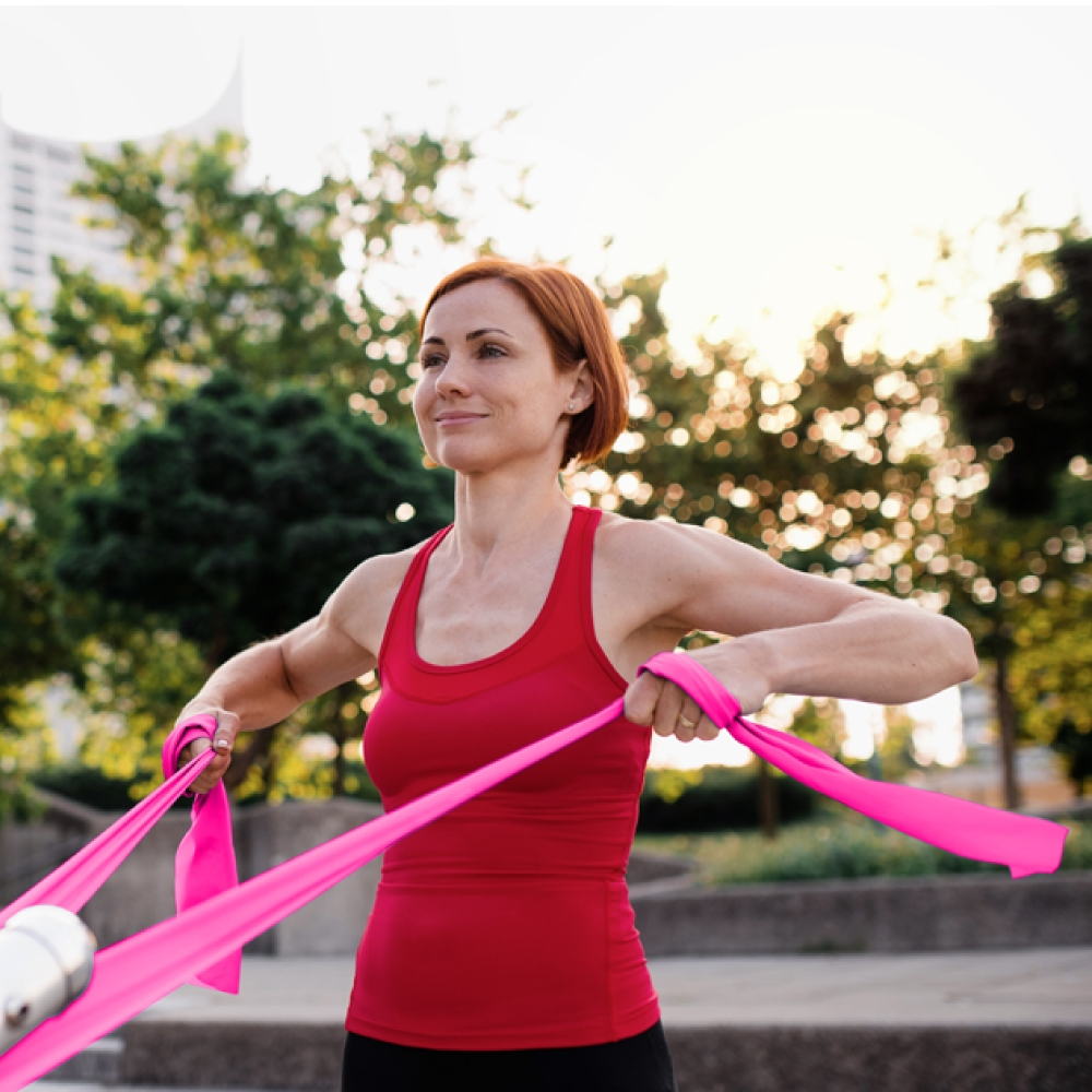 Mujer pelirroja entrenando con banda elástica rosa al aire libre.