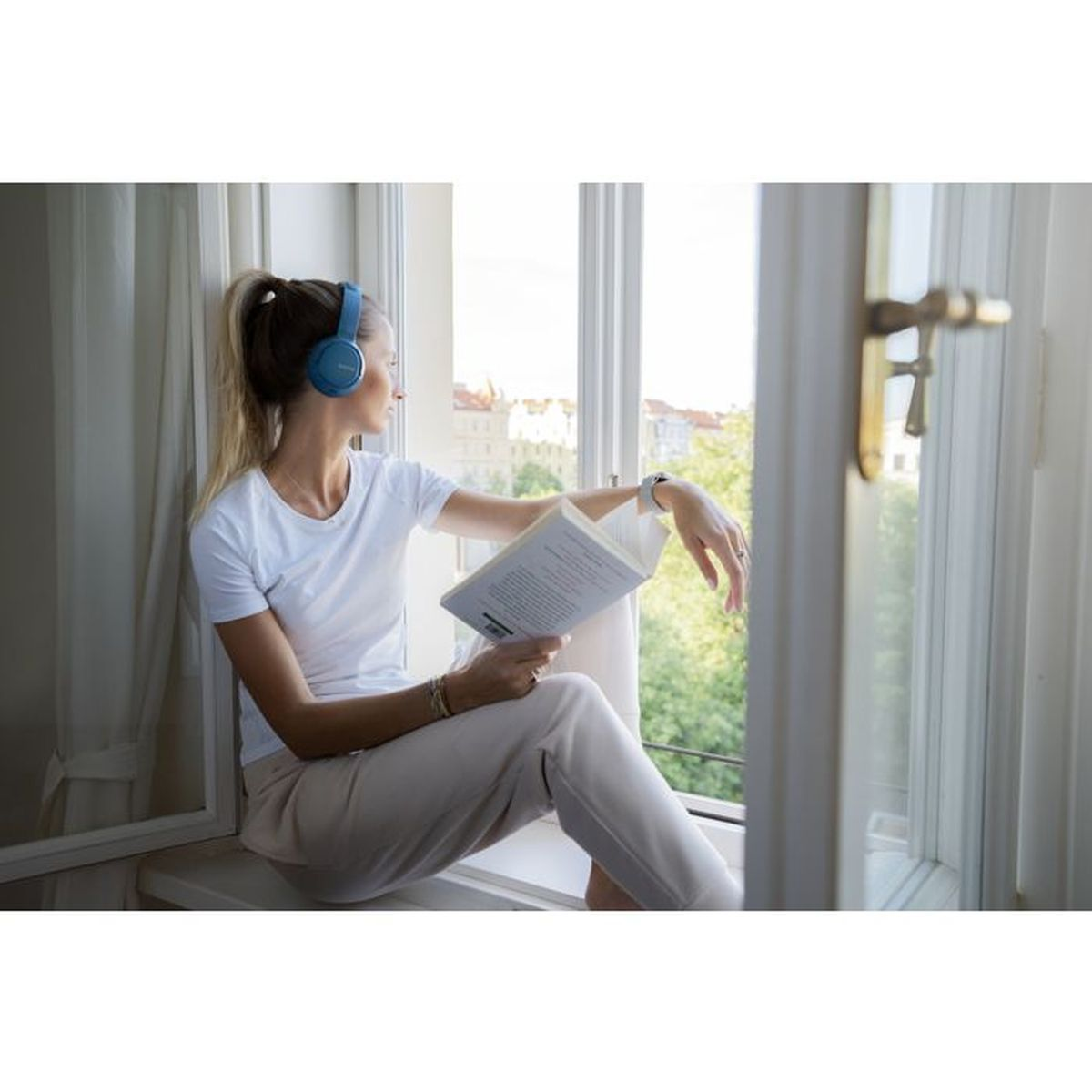 Donna con cuffie legge libro sul davanzale, guarda fuori.