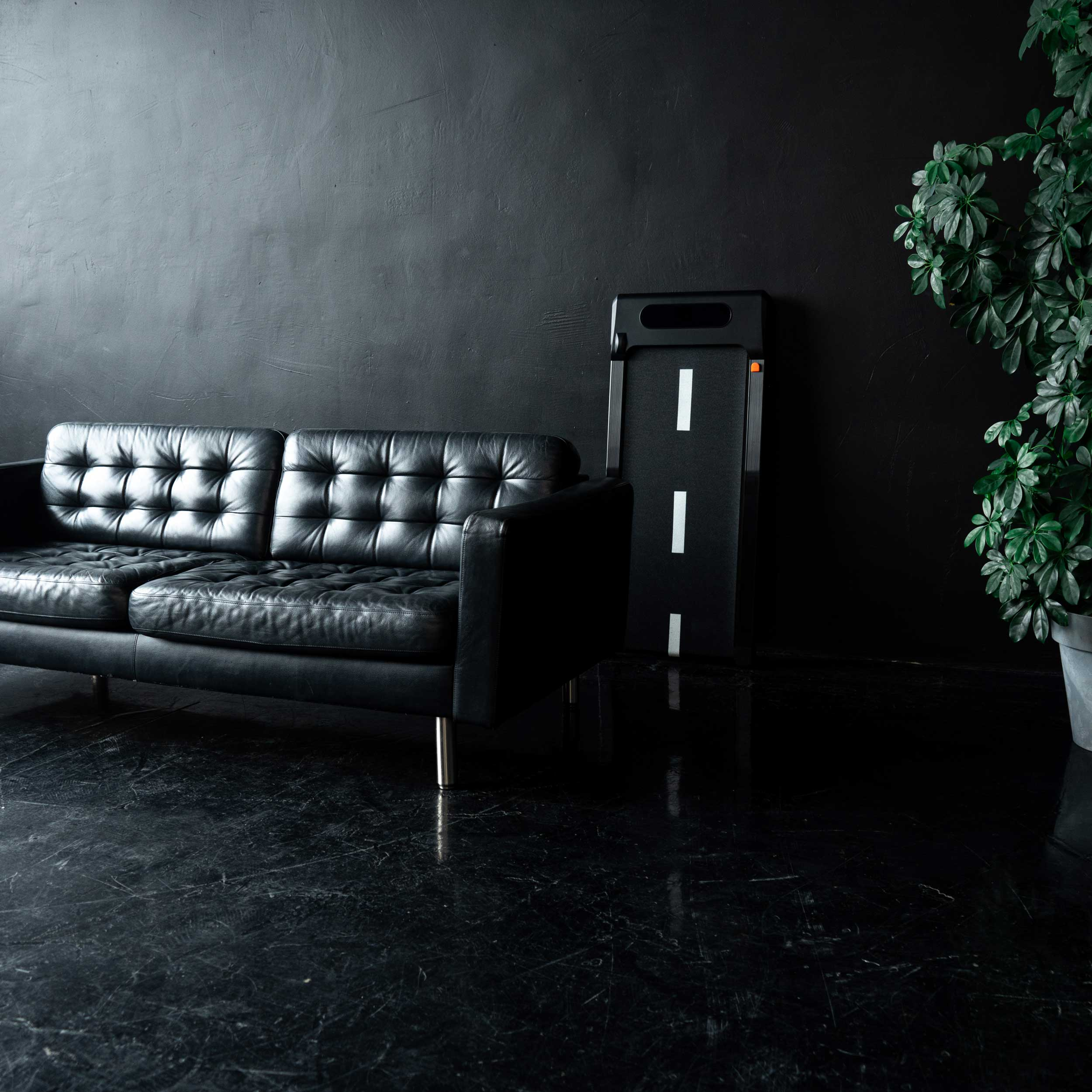 Czarna skórzana kanapa na ciemnej ścianie z rośliną doniczkową i bieżnią.