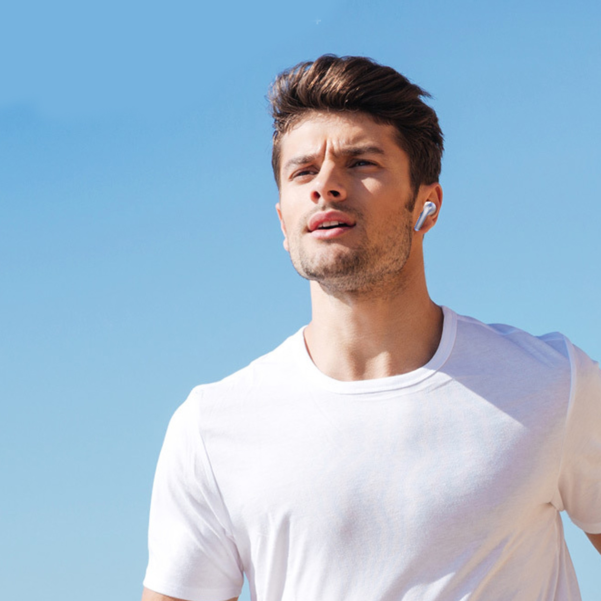 Ein Mann mit weißem T-Shirt und Ohrhörer, blauer Himmel.