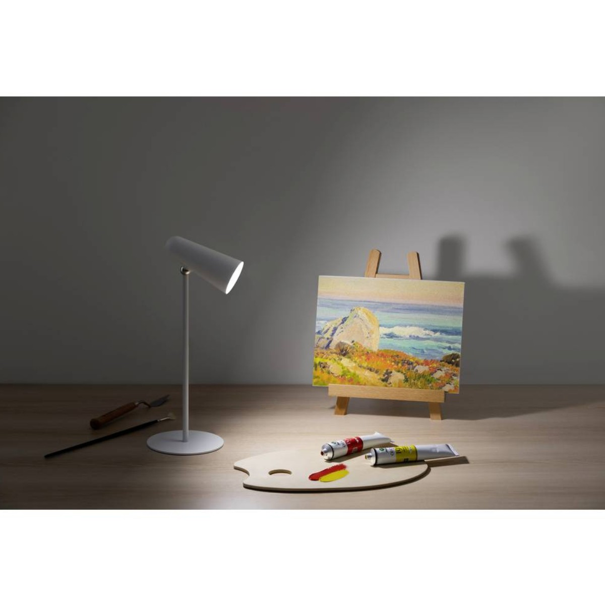 Een schilderij van een kustlandschap staat op een ezel. Een lamparm en verfpaletten rusten in de buurt.