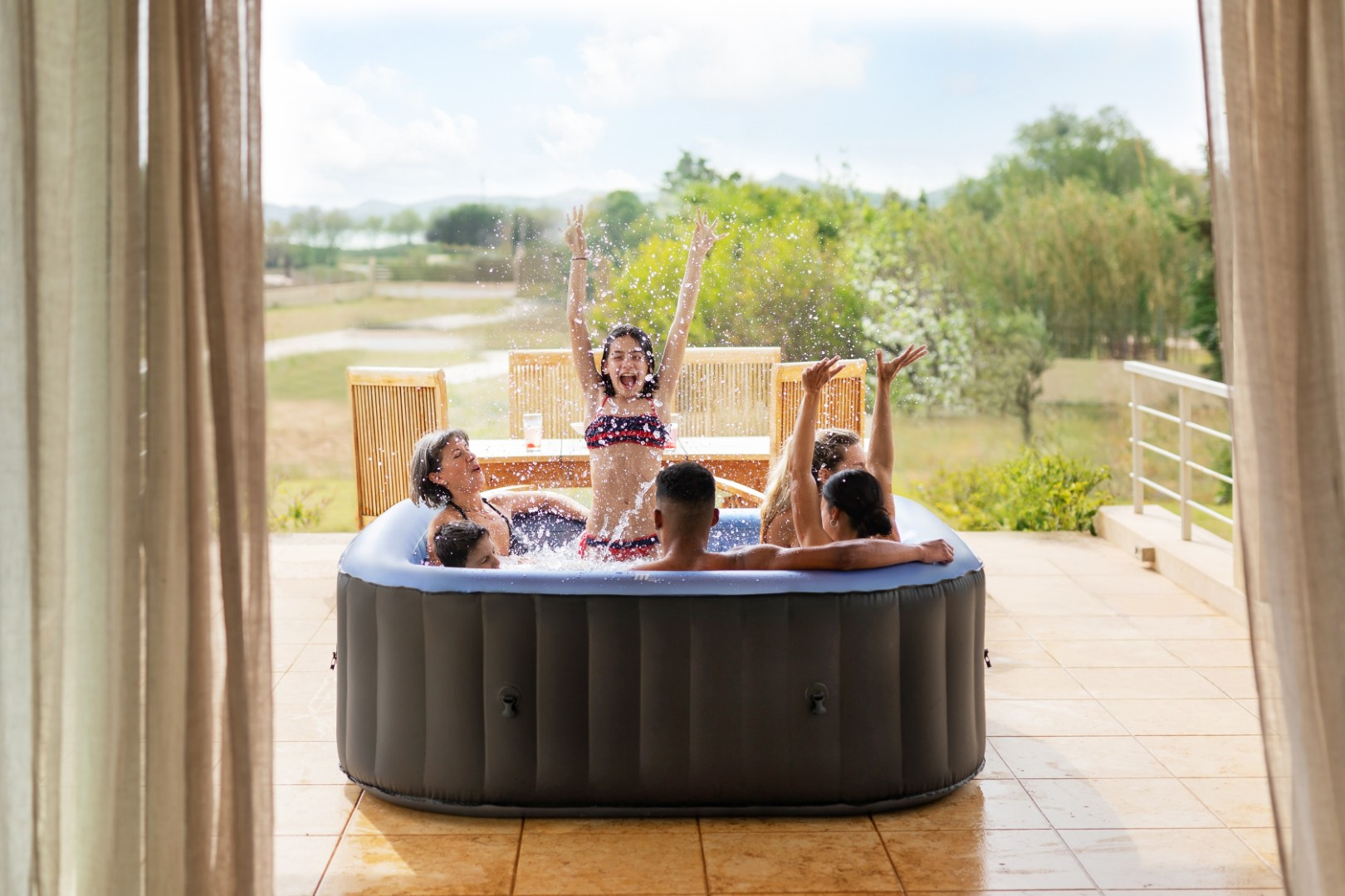 Menschen planschen in einem aufblasbaren Whirlpool auf einer Terrasse mit Blick auf eine grüne Landschaft.