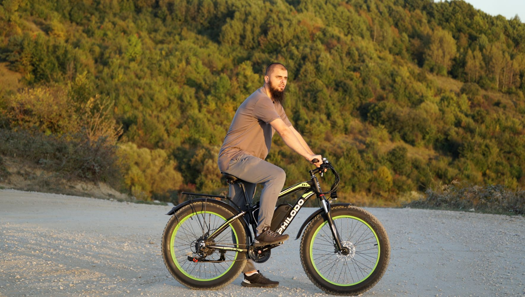 Uomo con barba guida e-bike con copertoni larghi su strada sterrata, collina boscosa.