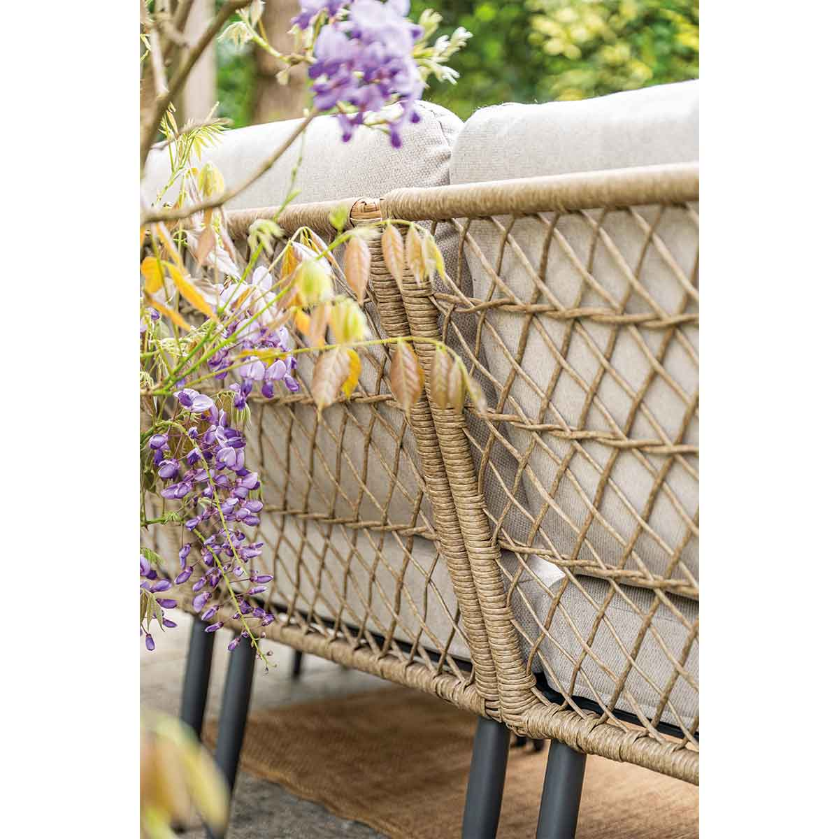 Un canapé en rotin avec des coussins gris, des fleurs violettes et des feuilles pendent à côté.
