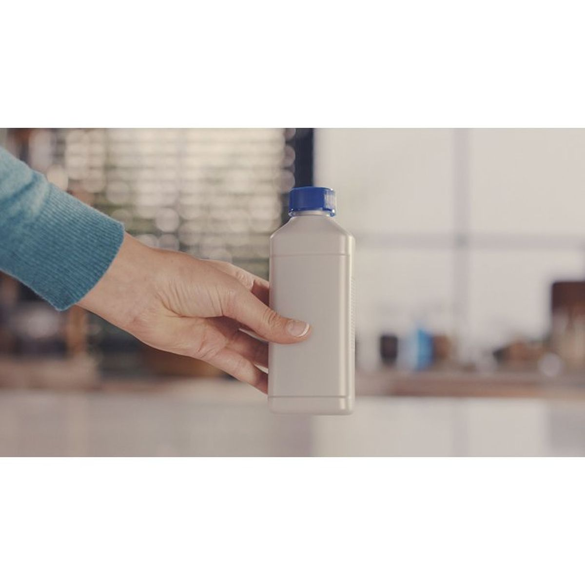 Eine Hand hält eine weiße Flasche mit blauem Deckel vor einem unscharfen Hintergrund.