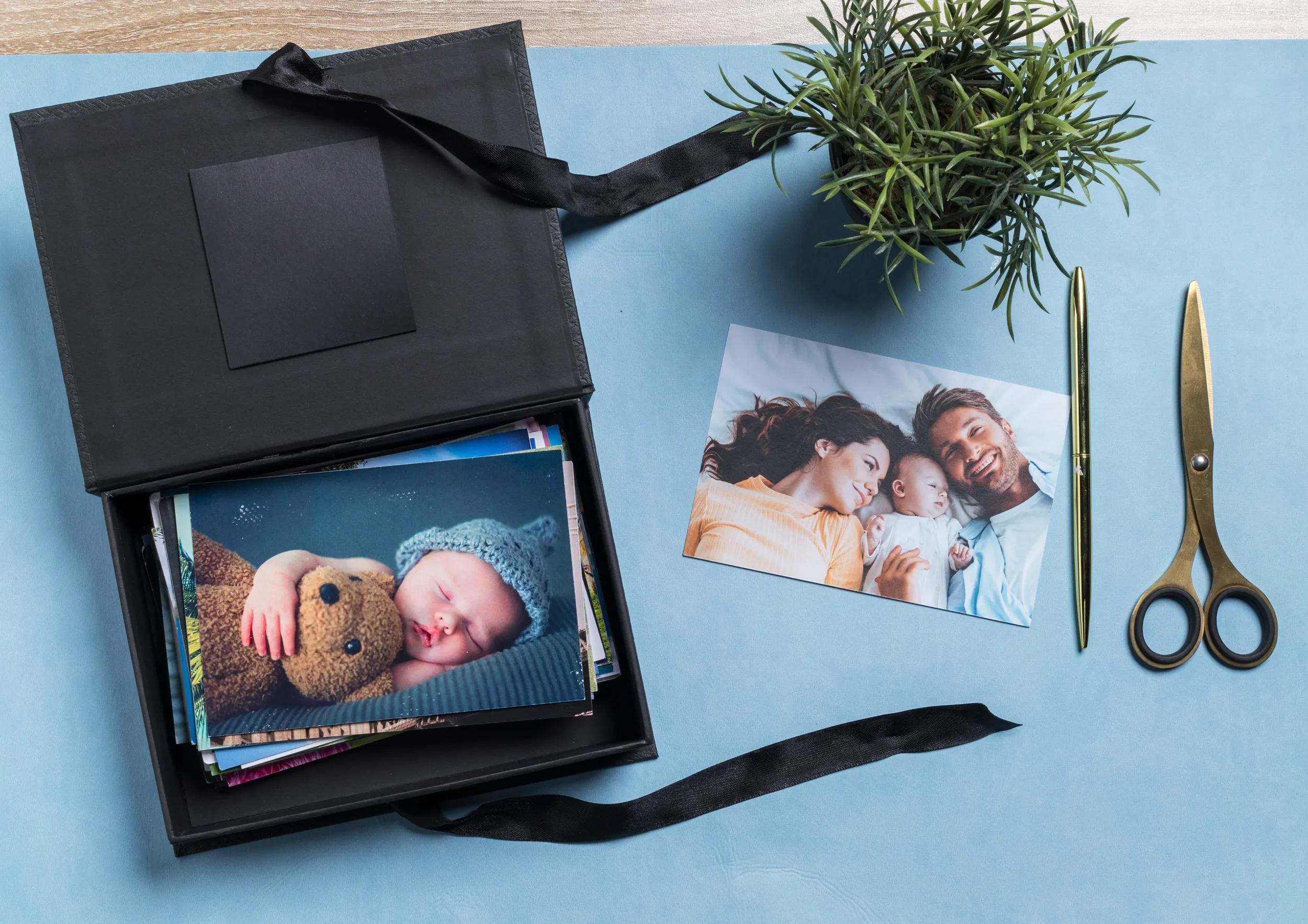 Caja negra con fotos, familia, bebé, planta, bolígrafo y tijeras sobre fondo azul.