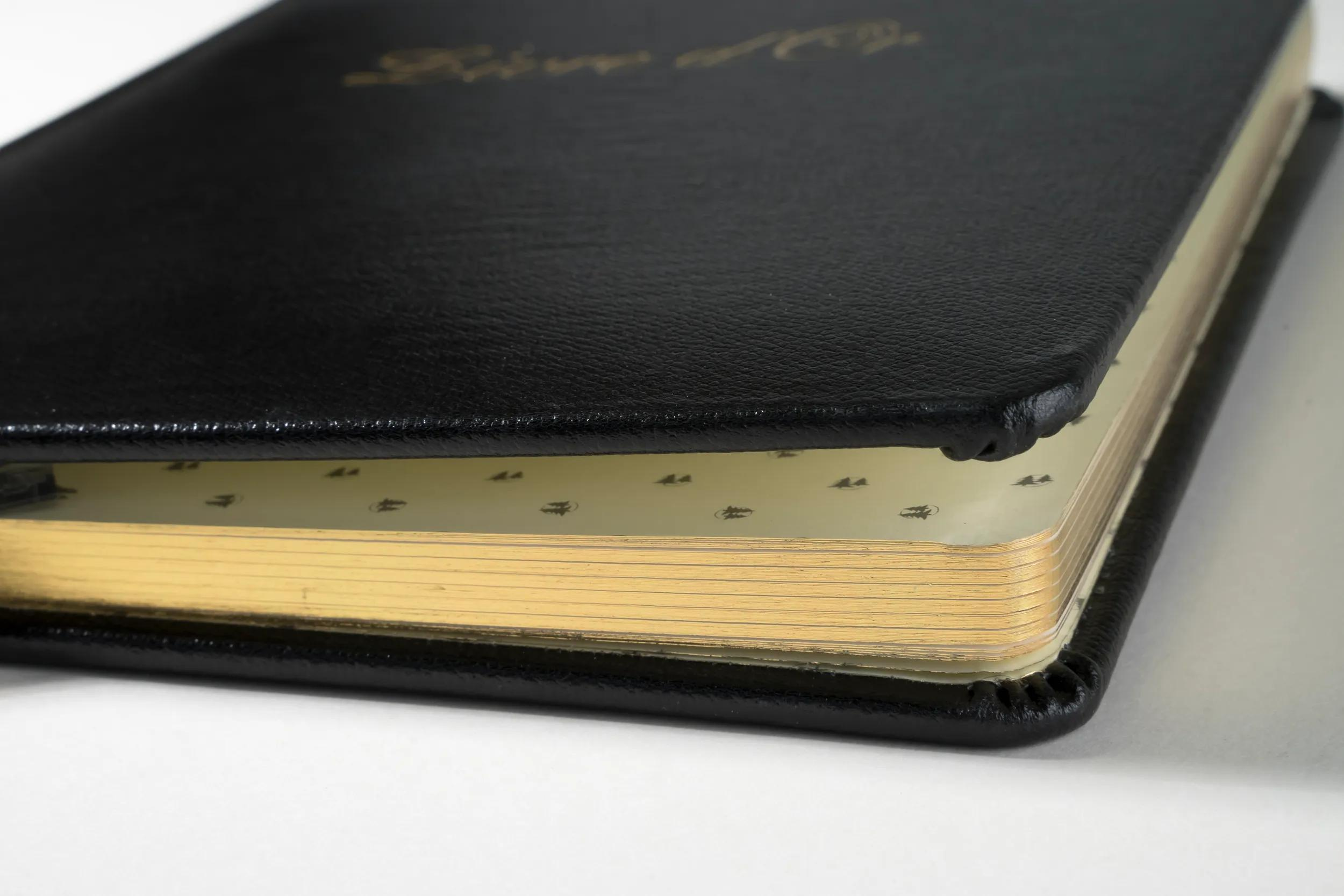 Un libro de cuero negro con páginas de borde dorado sobre superficie blanca.