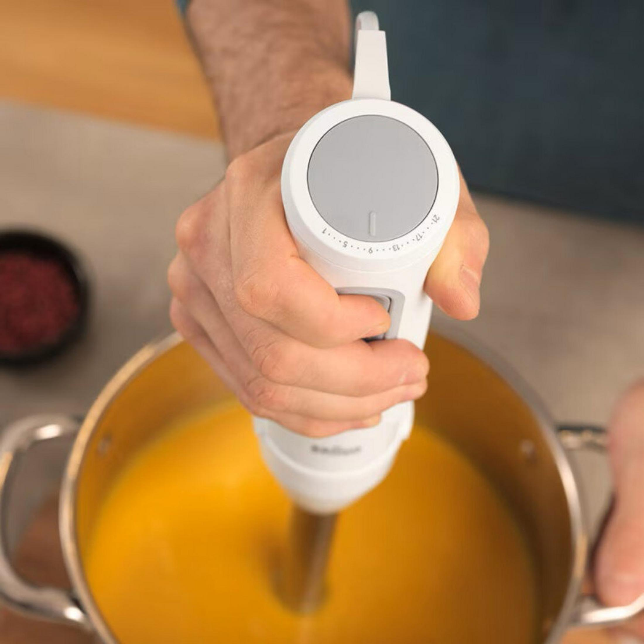 Een hand gebruikt een witte staafmixer in een pan met gele soep.